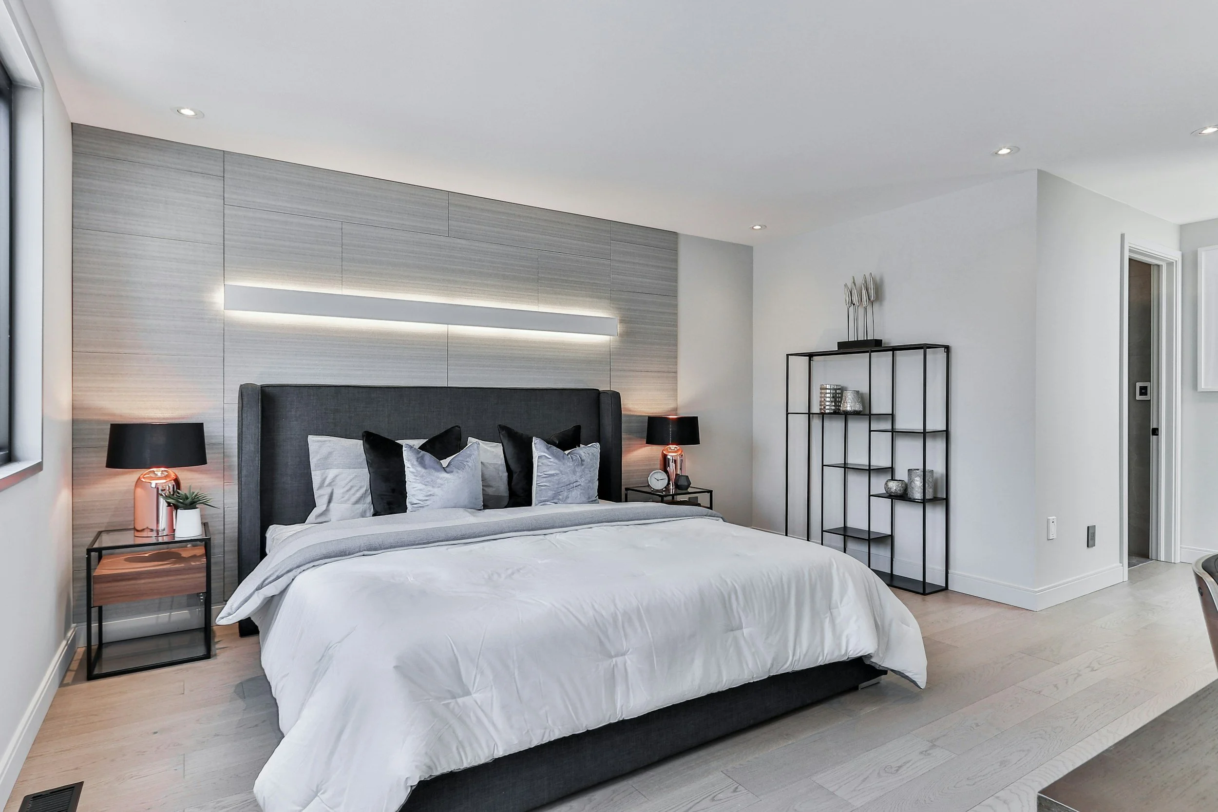 Modern bedroom with a gray upholstered bed, black side tables with matching lamps, a black metal shelving unit, white walls, and light wooden flooring.