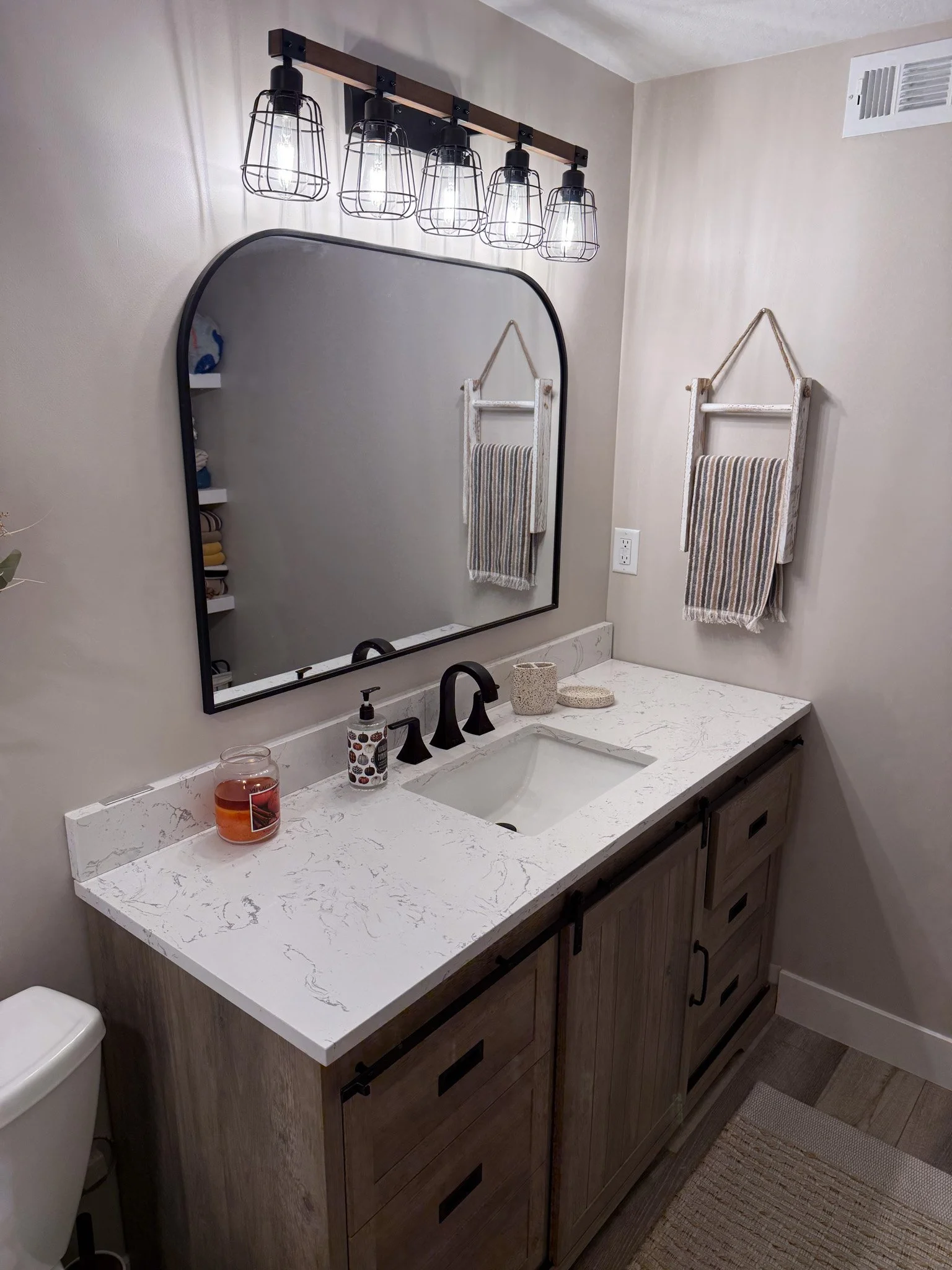 Bathroom vanity with a marble countertop, black faucet, large mirror, wall-mounted sconce with exposed bulbs, striped hand towels, and decorative items.