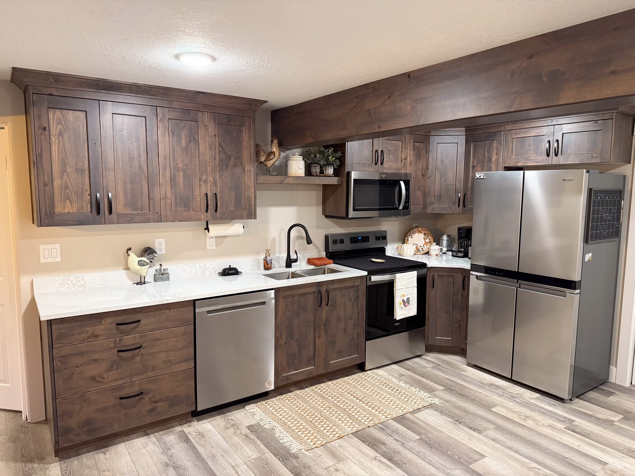 Kitchen with dark wooden cabinets, white countertop, stainless steel appliances including a refrigerator, microwave, stove, and dishwasher. There are decorative items on the countertop and upper shelf, and a beige rug on wood-look flooring.