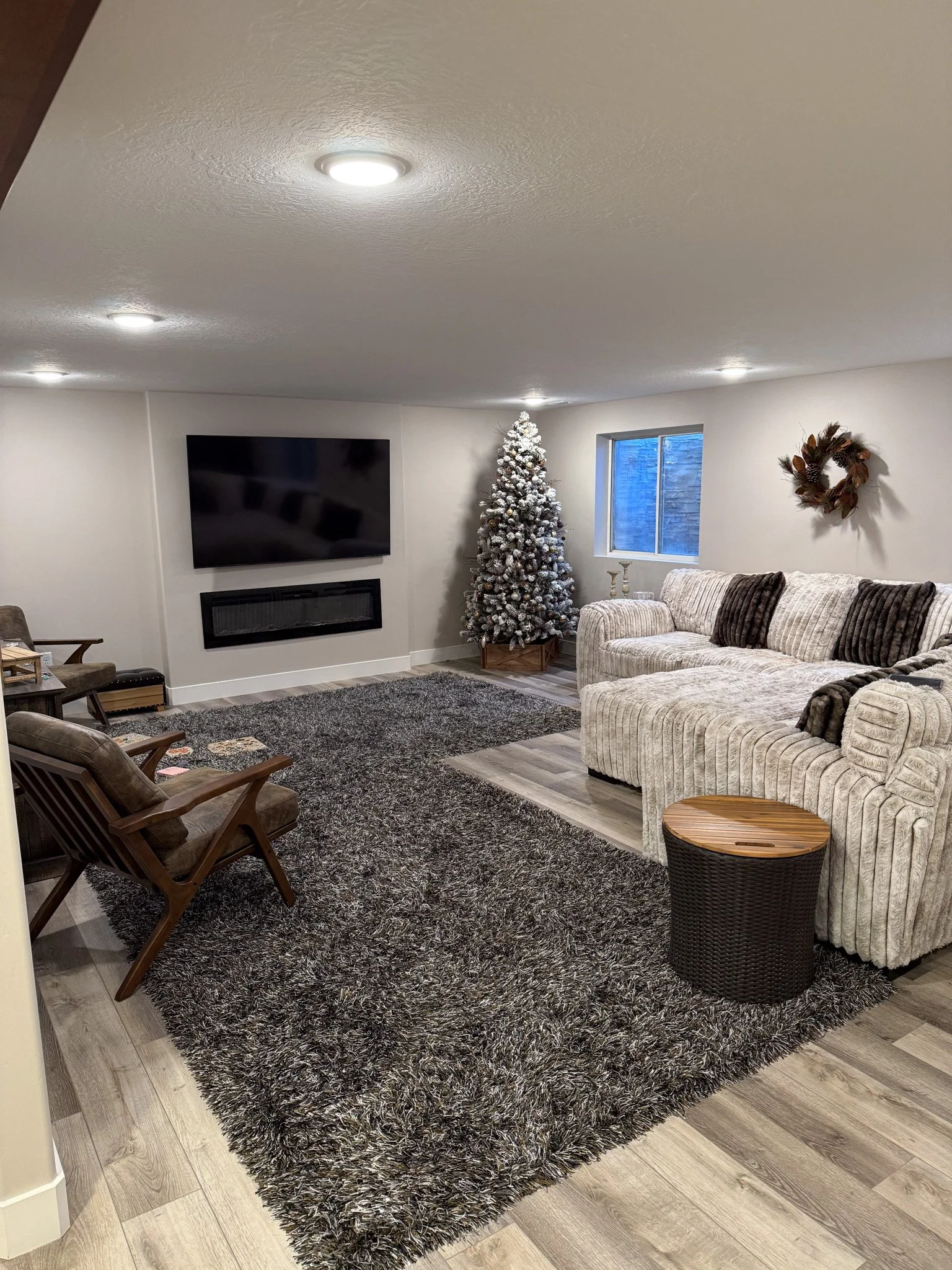 Living room with a large white sofa, a Christmas tree with snow-flocked branches, a wall-mounted television above a fireplace, two wooden chairs, a dark shaggy area rug, a small circular side table, a window, and a holiday wreath on the wall.