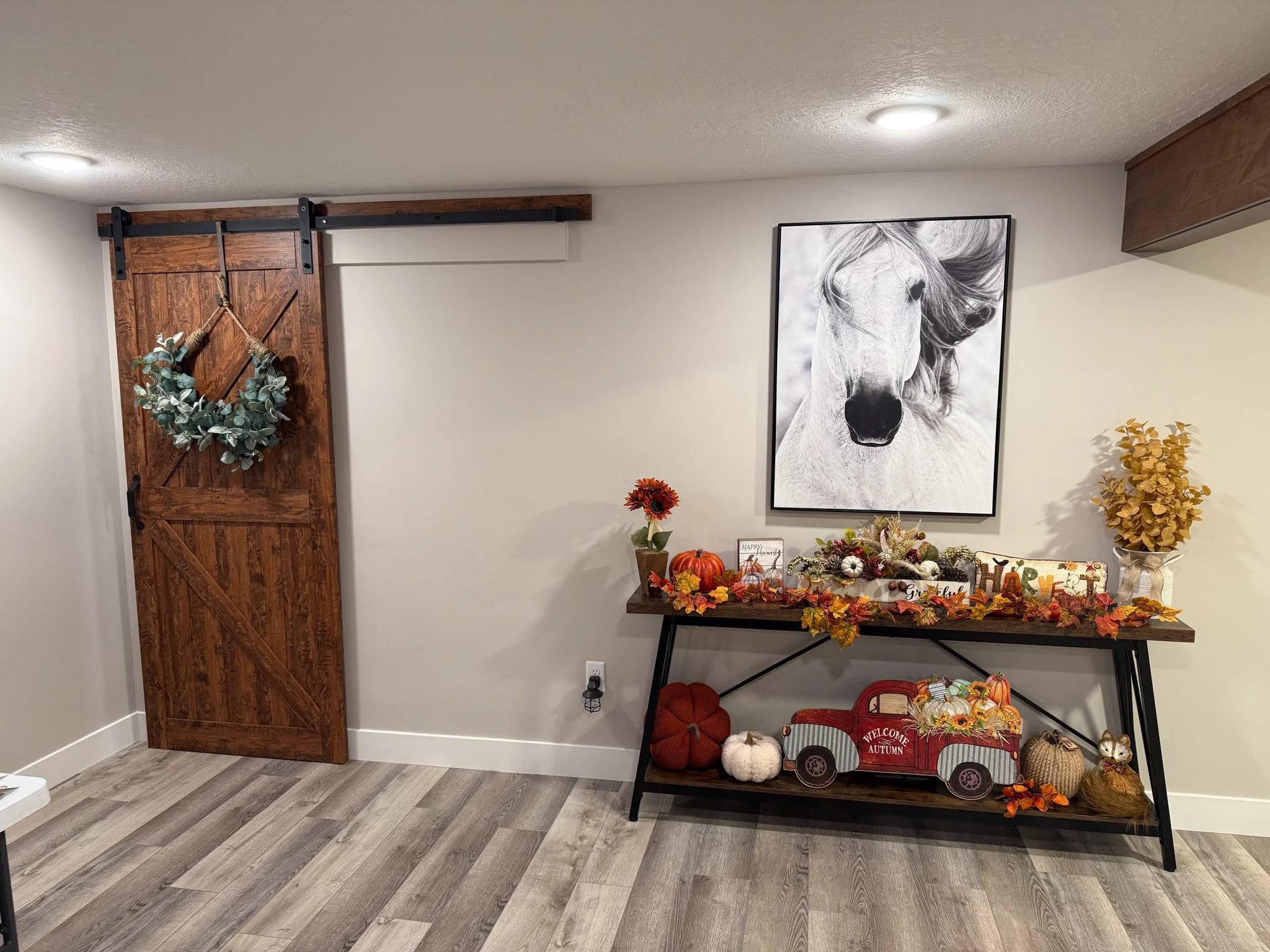 Interior of a room with a decorative fall-themed display. There is a wooden sliding barn door with a eucalyptus wreath, a black and white horse poster on the wall, and a black console table decorated with pumpkins, flowers, and autumn leaves. The flo