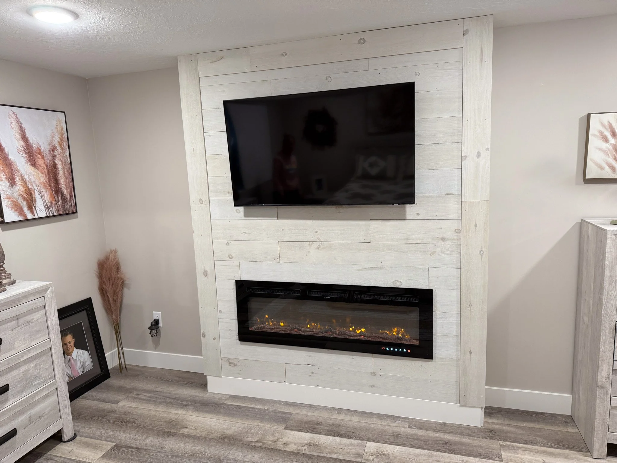 Living room with white shiplap feature wall, mounted flat-screen TV, and electric fireplace, framed pictures, and decor on side cabinets, with light wood flooring.