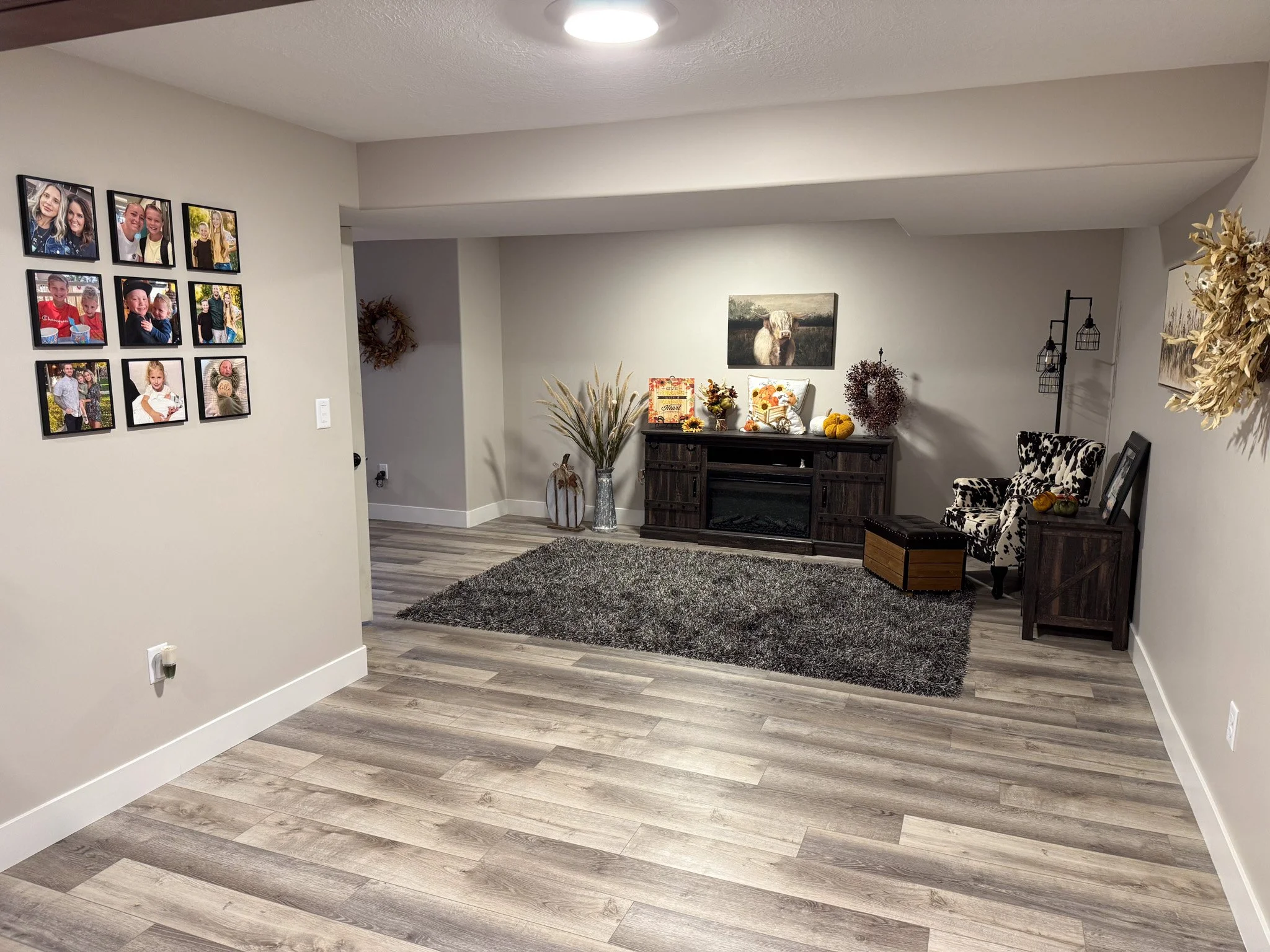 A cozy living room with a gray rug, a black fireplace, a dark wood console table decorated with fall harvest decor, a black and white patterned armchair, framed family photos on the wall, and decorative plants and artwork, with wood laminate flooring