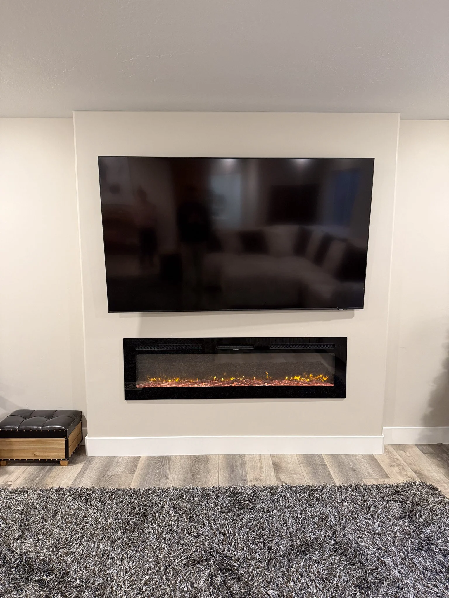 Flat screen television mounted on a wall above a modern electric fireplace with glowing flames, with a gray shaggy rug in front.