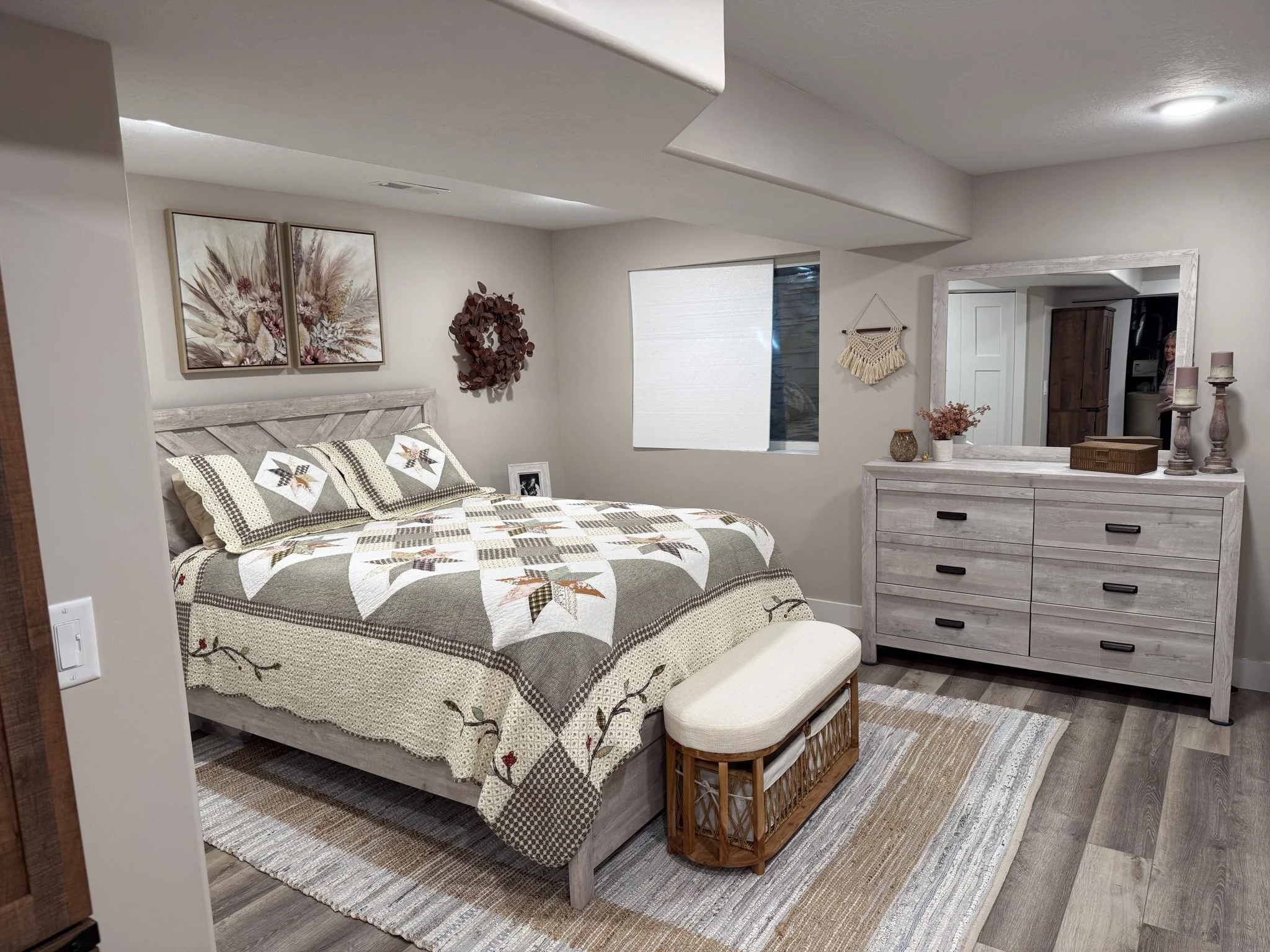 A cozy bedroom with a queen-sized bed featuring a patchwork quilt and matching pillows, a white wooden headboard, framed botanical artwork, a wall wreath, a large window with a rolled-up shade, a white dresser with decor items, a mirror, a bench at t