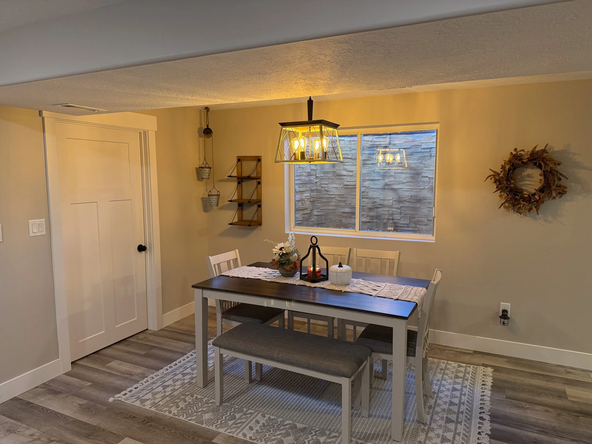 Dining room with a wooden table, four chairs, a bench, a hanging light fixture, a window, wall decor, and a rug on the floor.