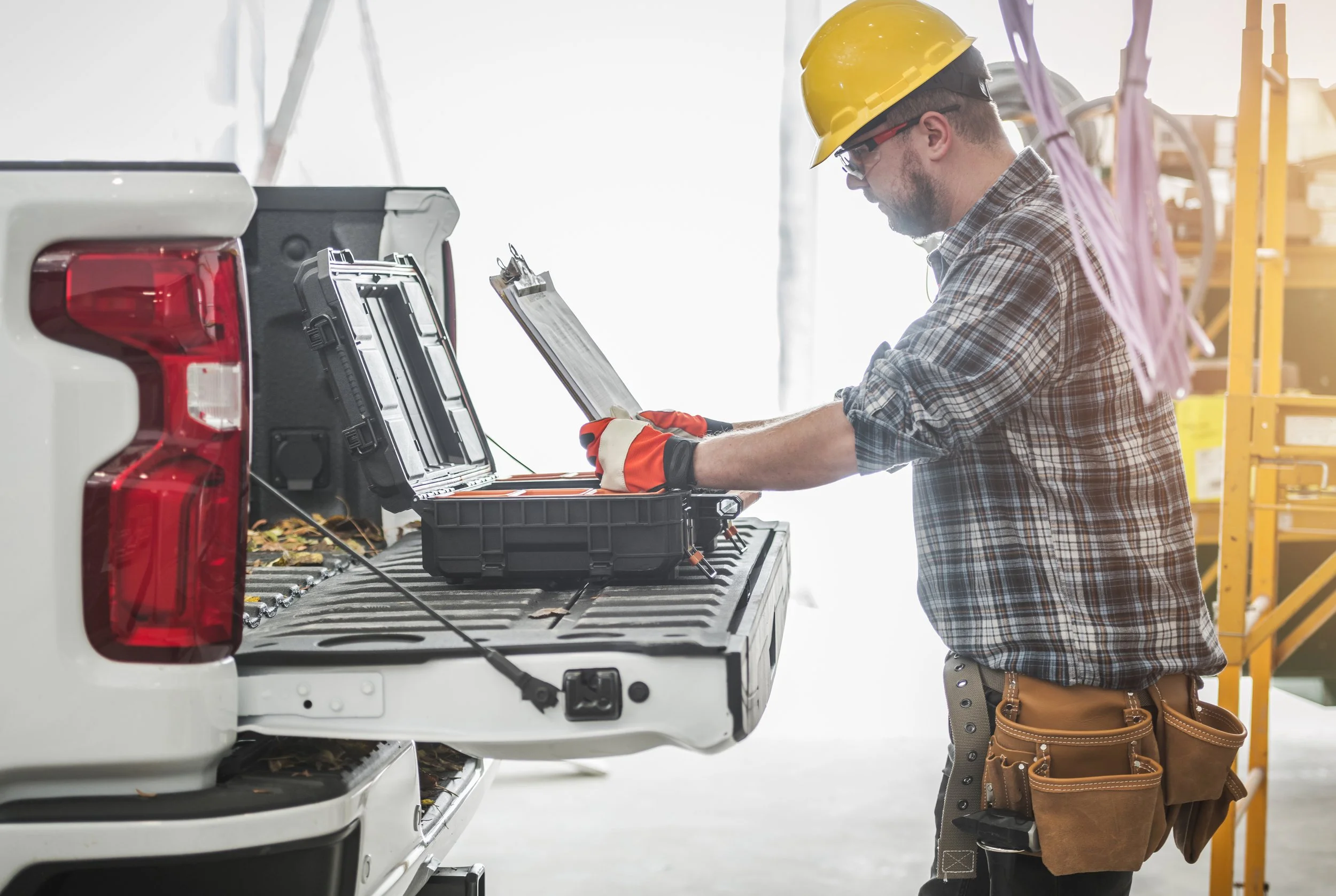 A man wearing safety glasses, a yellow hard hat, gloves, a plaid shirt, and a tool belt working with electronic equipment in the back of a white truck inside an industrial or warehouse setting.