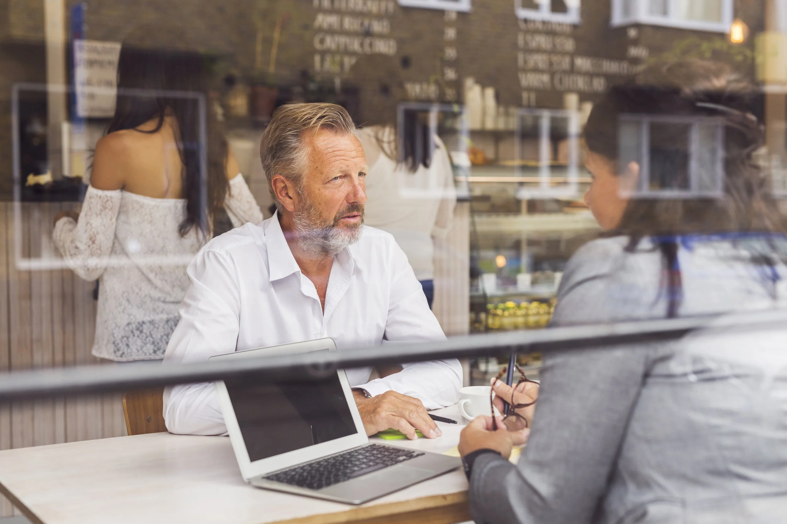 Two people having a conversation in a coffee shop, viewed through a glass window with reflections. One is a man with gray hair and a beard, wearing a white shirt, sitting at a table with a laptop and a cup. The other is a woman with dark hair, facing him, holding glasses.