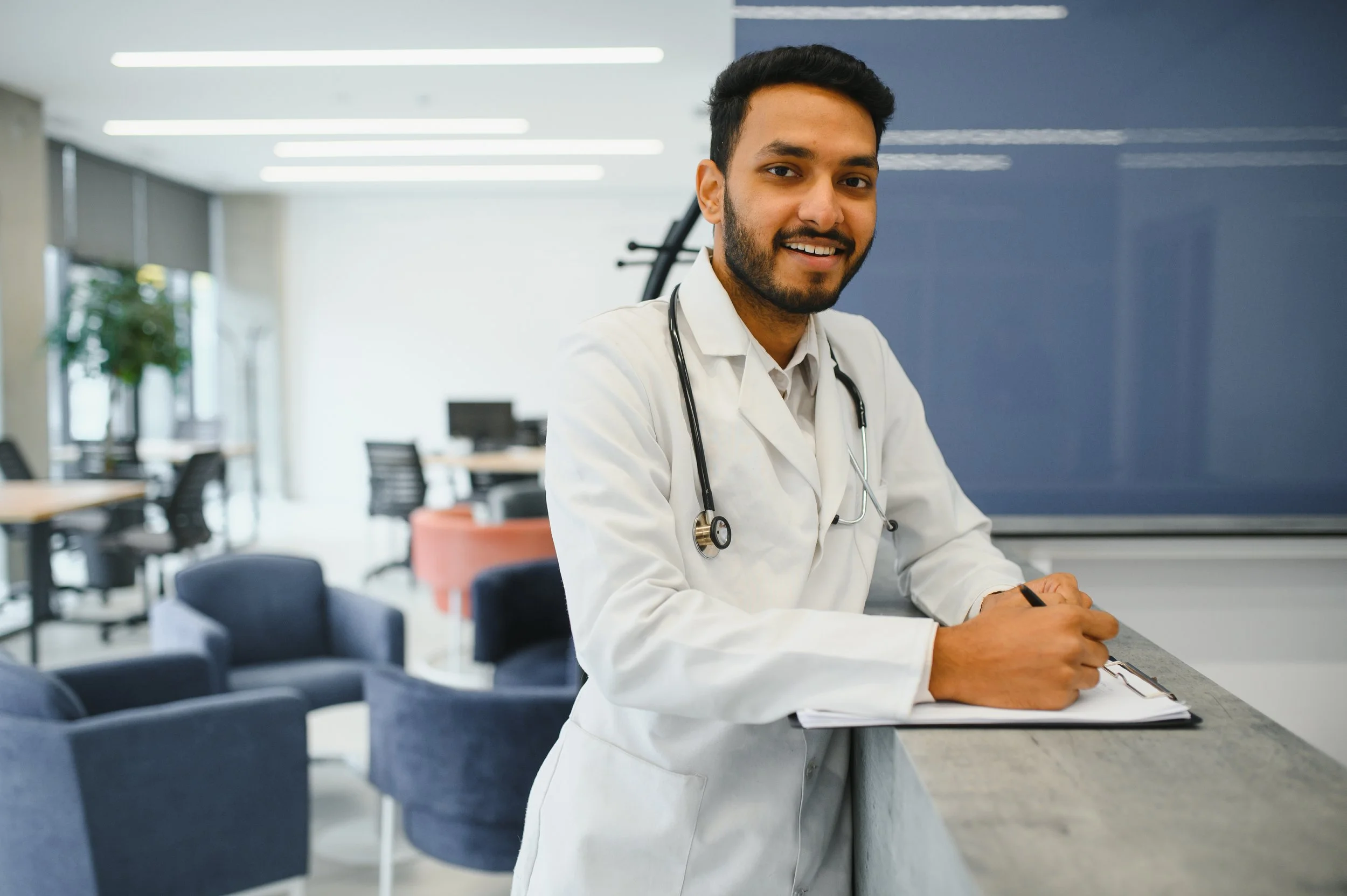 Smiling male doctor with a stethoscope around his neck, writing on a clipboard in a modern office or medical facility.