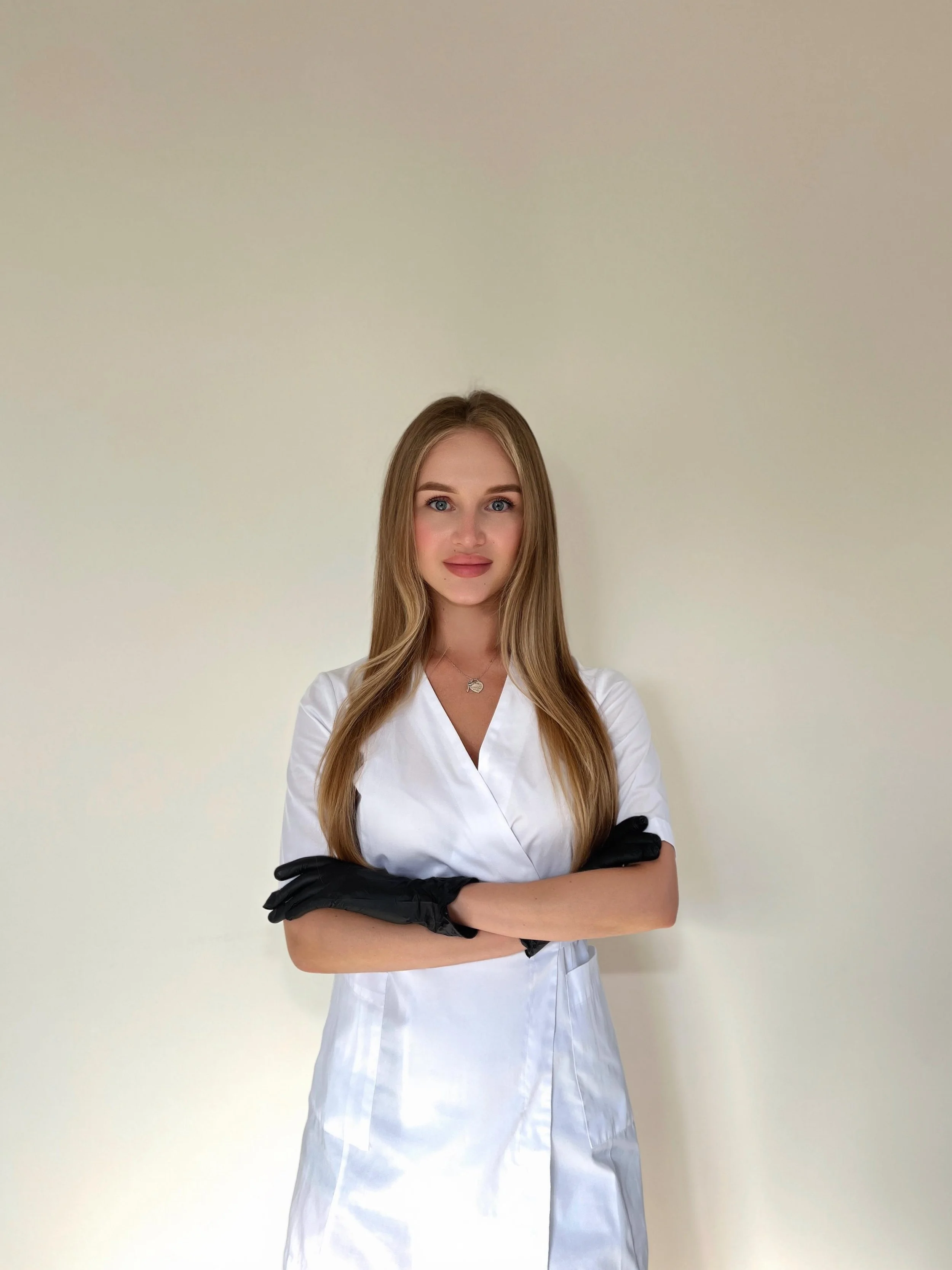 A woman with long blonde hair wearing a white medical coat and black gloves, standing with arms crossed against a plain off-white background.