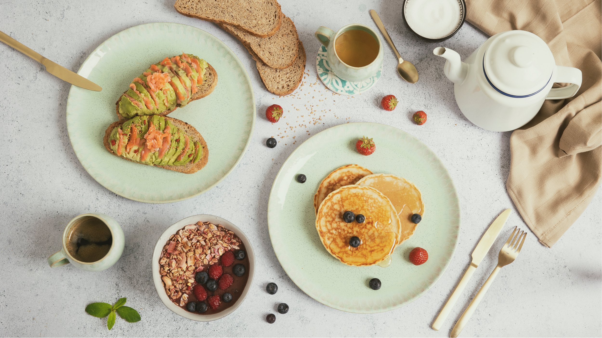 Petit déjeuner avec des pancakes aux myrtilles, des toasts à l'avocat, un bol de granola avec des fruits, une théière, des tasses, des fruits frais, et des ustensiles sur une table claire.