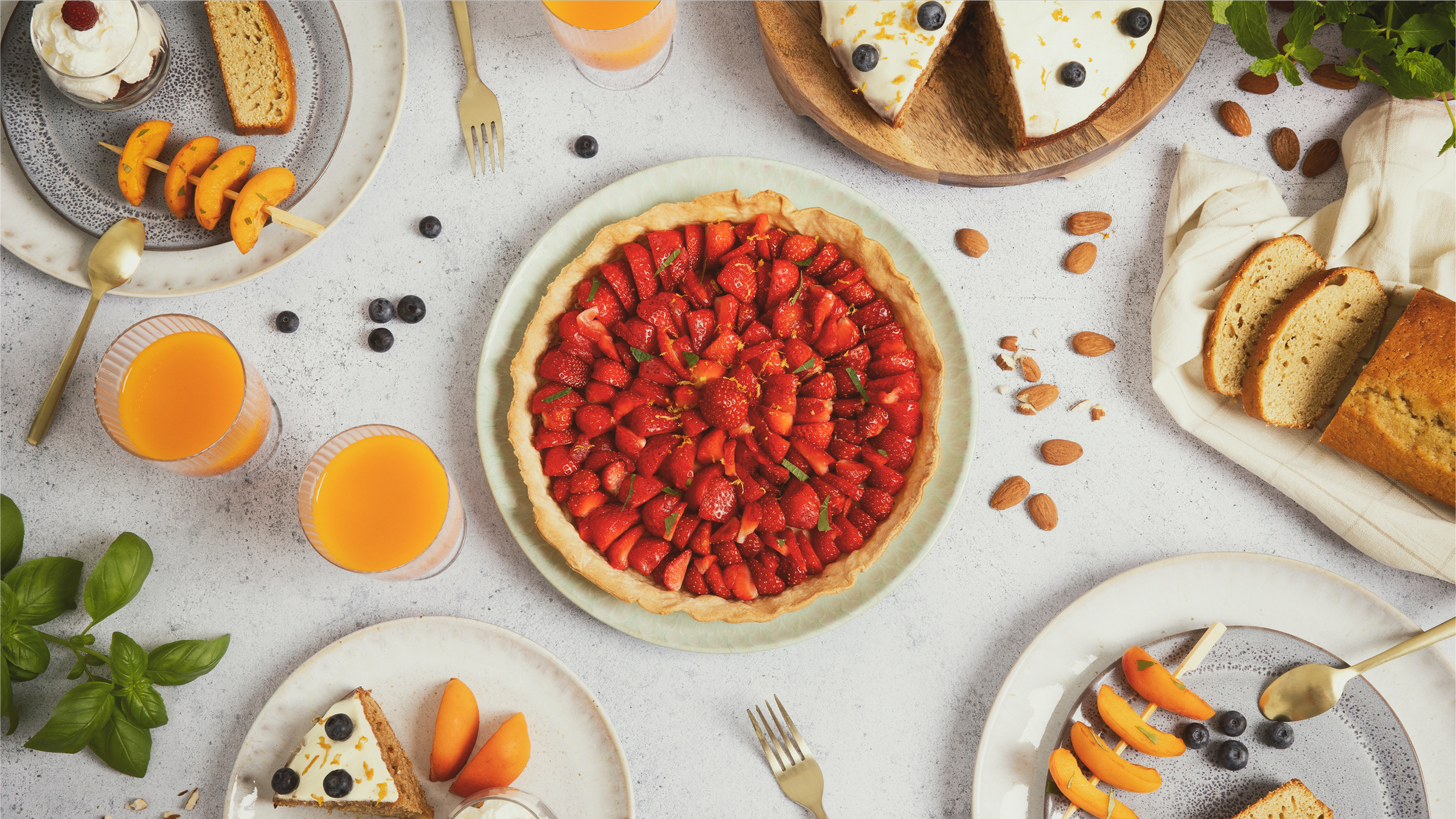 Gâteau aux fraises au centre, tranches de cake, tartes, jus d'orange, fruits comme myrtilles, abricots, et amandes sur la table.