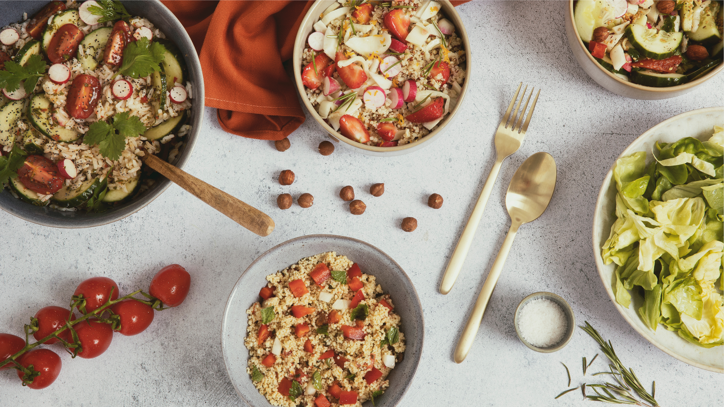Assortiment de salades et de tomates cerises sur une table, avec des couverts dorés et des herbes aromatiques.