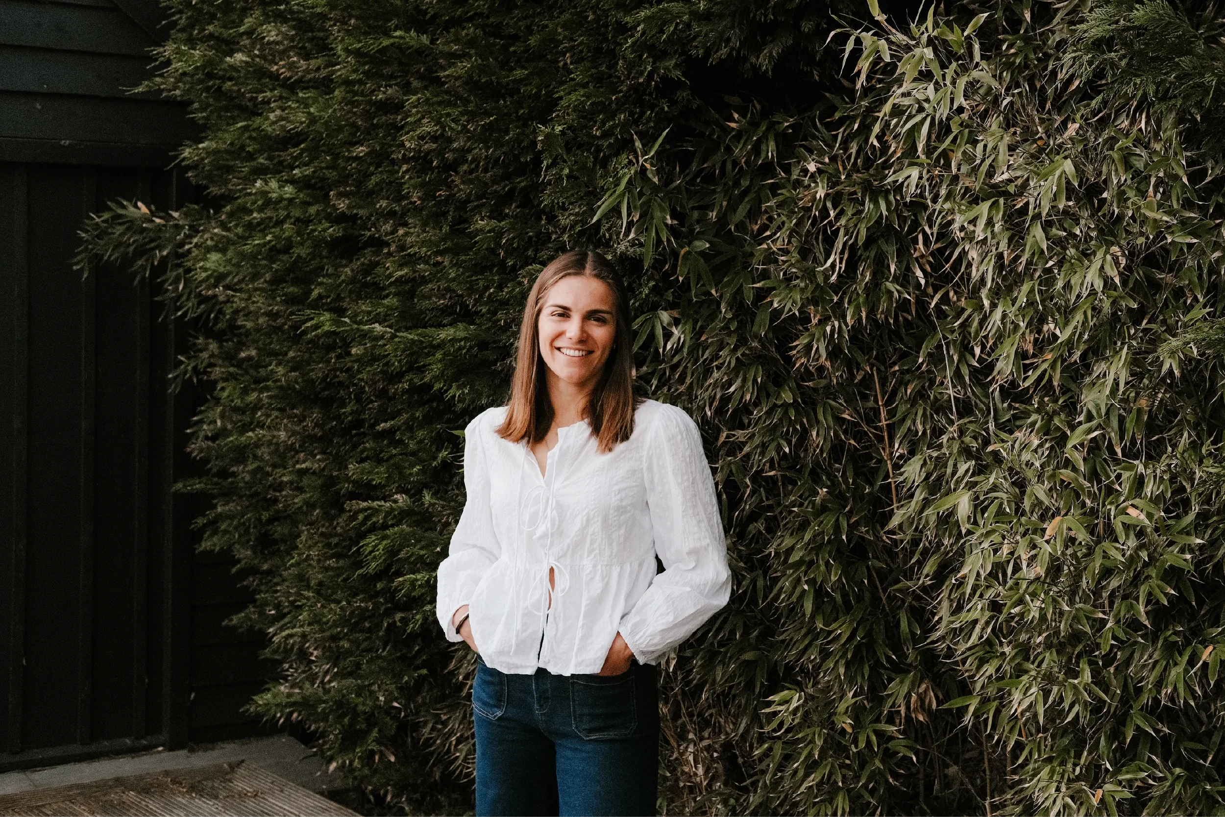Portrait of Georgina Howell standing in front of a hedge with her hands in her pockets, smiling