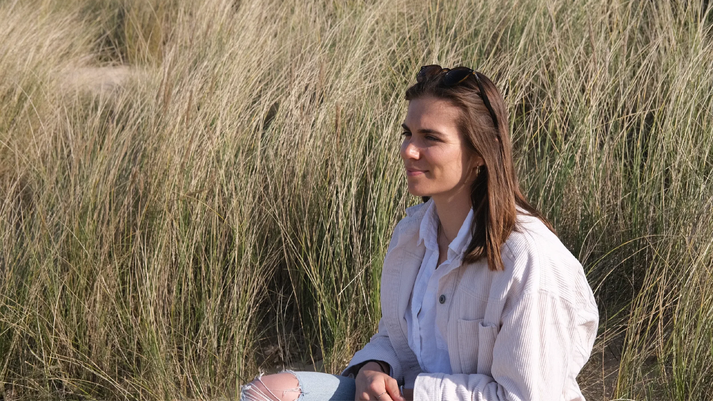 Side-on portrait of Georgina Howell sitting in tall grass at the beach.