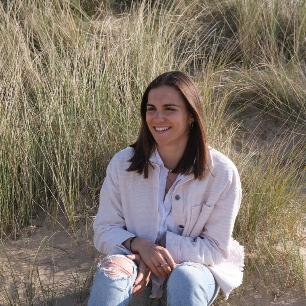 Portrait of Georgina Howell sitting on the sand at the beach with tall grass in the background, smiling and looking to her right, wearing a white jacket and ripped jeans.