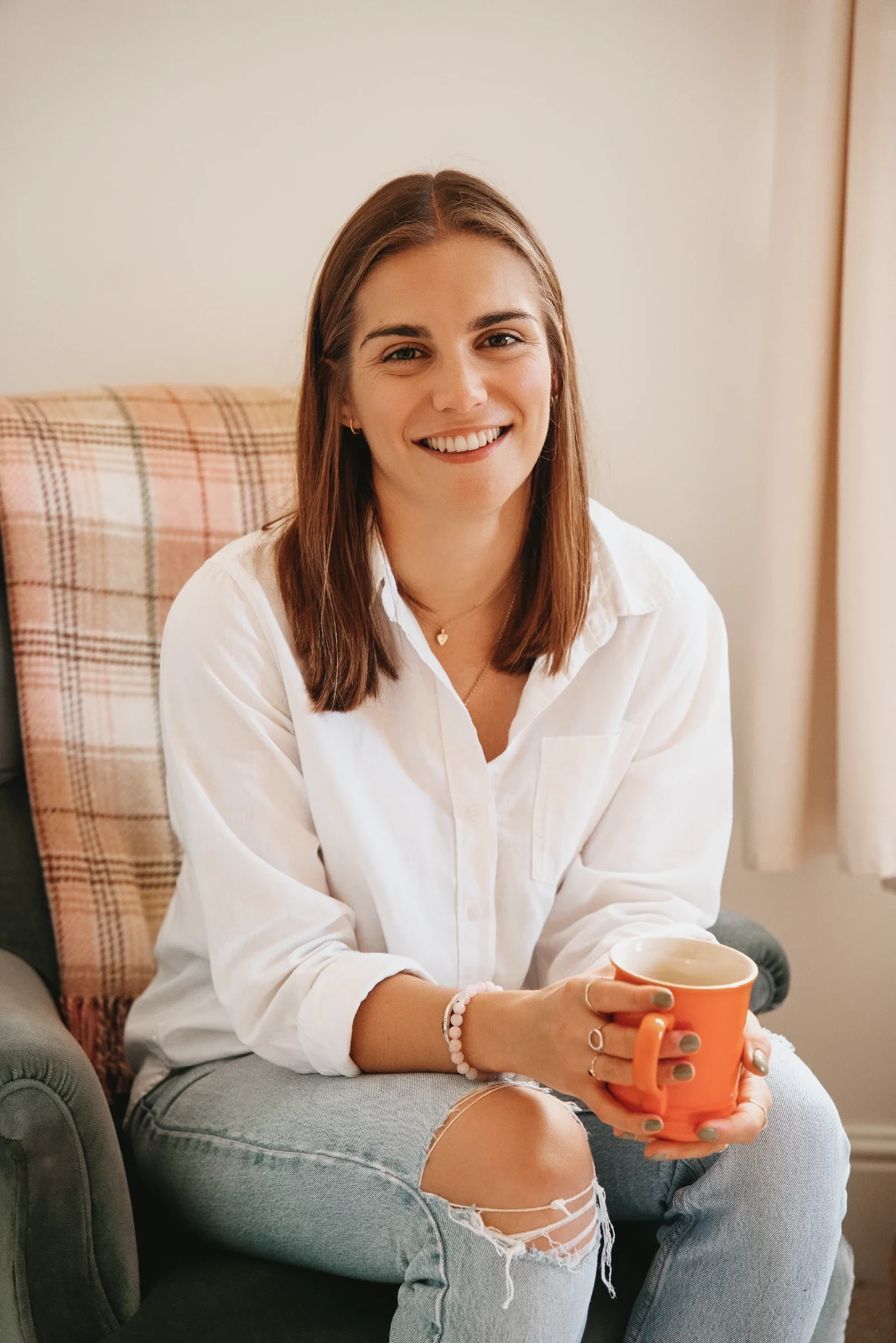 Portrait of Georgina Howell sitting on armchair, smiling, holding orange mug.
