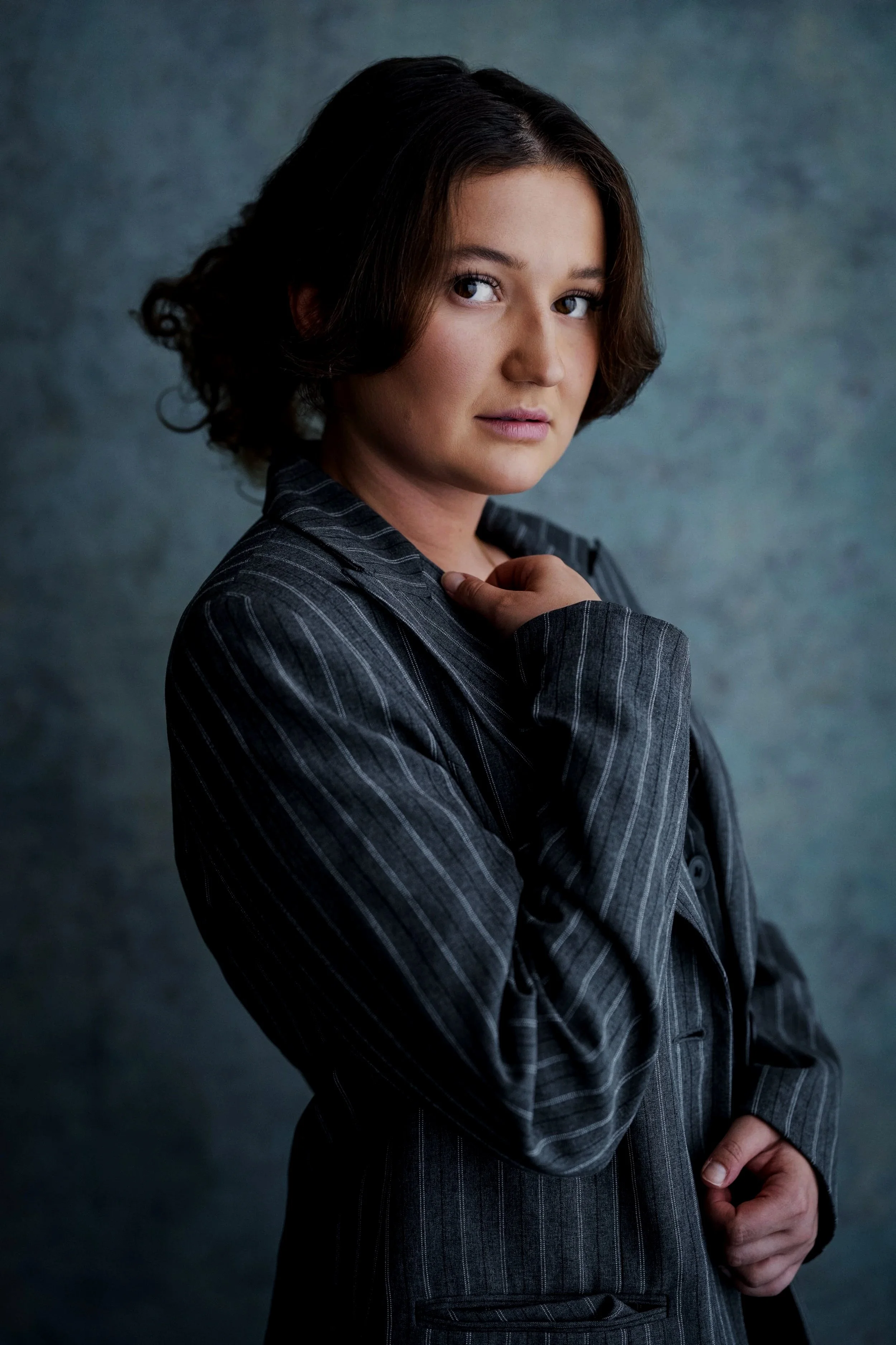 A woman with short, wavy brown hair and light skin wearing a dark pinstripe suit, looking at the camera with a slight smile, against a textured blue background.