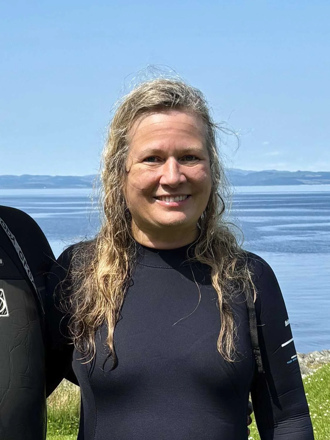 A woman with long blond hair smiling outdoors near a body of water with hills in the distance.