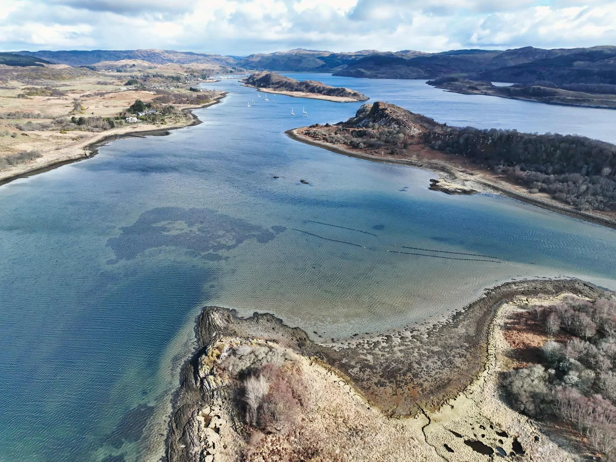 Aerial view of a river with boats, surrounding hills with sparse trees, and a rocky shoreline.