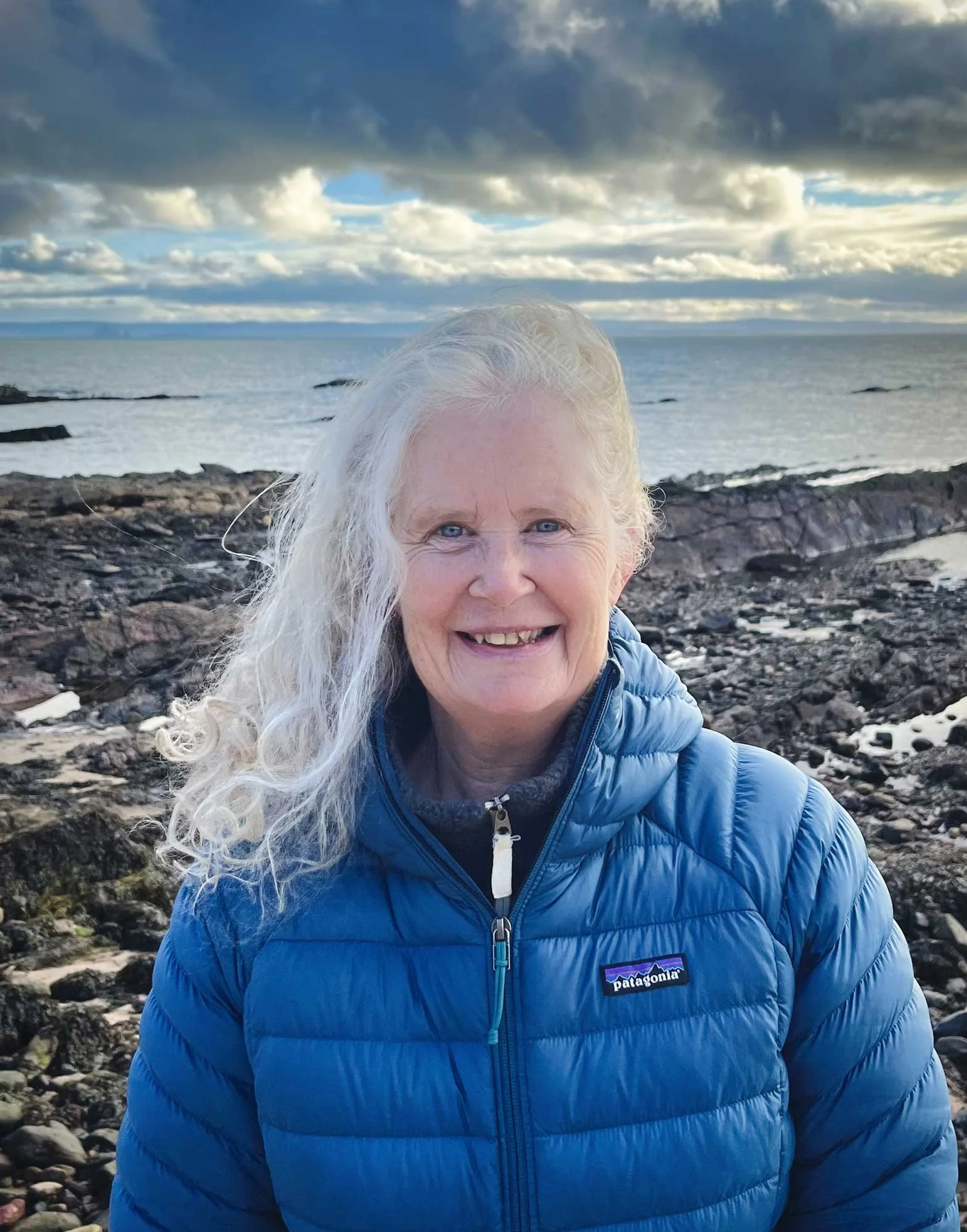 Smiling woman with long curly gray hair wearing a blue puffer jacket outdoors near the rocky shoreline and ocean with cloudy sky.