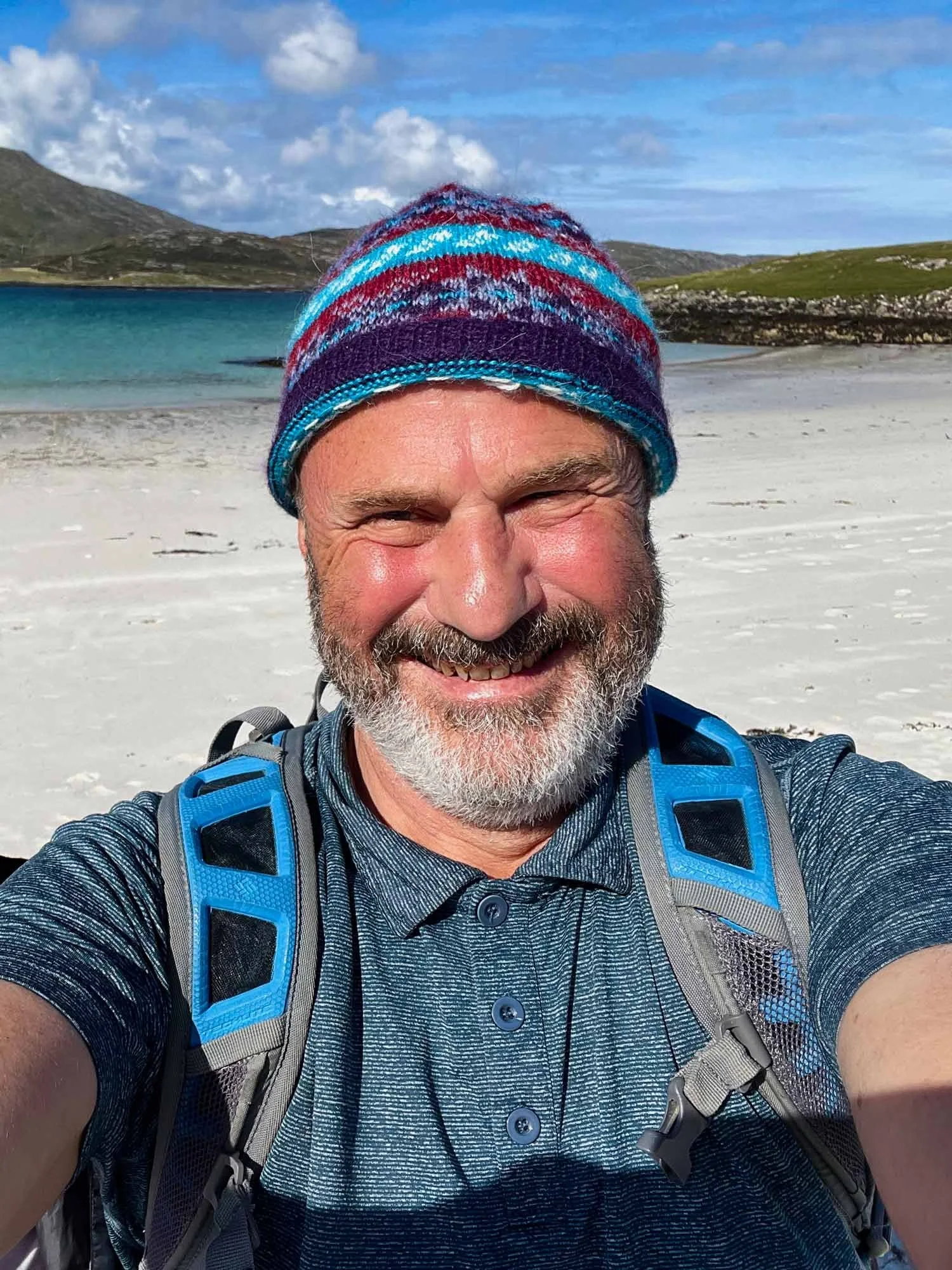 Pete standing on a Scottish shoreline with mountains and a blue sky in the background. Pete is smiling and wearing a knitted hat and rucksack. The sun is shining on his face.