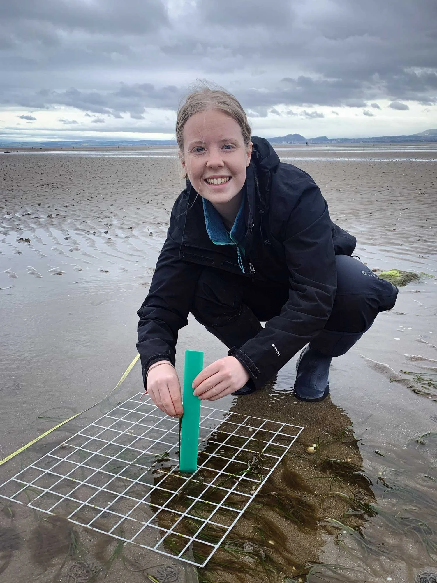 A woman on a beach, squatting near the water, holding a green tube and a grid, with seaweed and sand around her, overcast sky in the background.