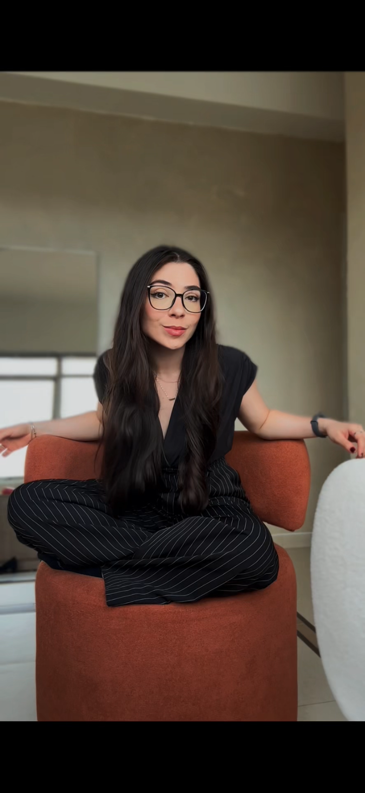 Mulher com longos cabelos castanhos escuros, óculos grandes, vestindo roupa preta com listras brancas, sentada de pernas cruzadas em uma poltrona laranja, com fundo de parede bege