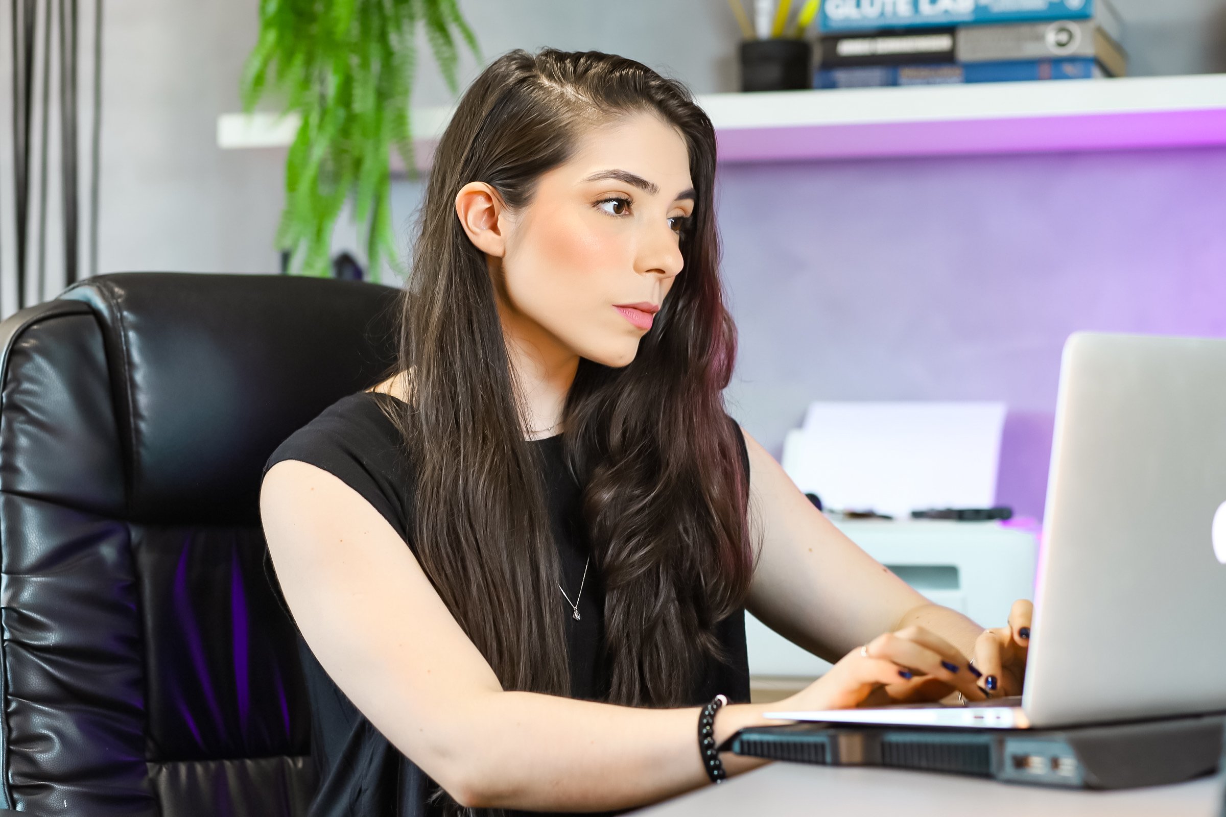 Mulher sentada em uma mesa de escritório usando um laptop, com cabelo castanho escuro, usando uma blusa preta e uma pulseira, com uma estante de livros ao fundo e uma planta ao lado.