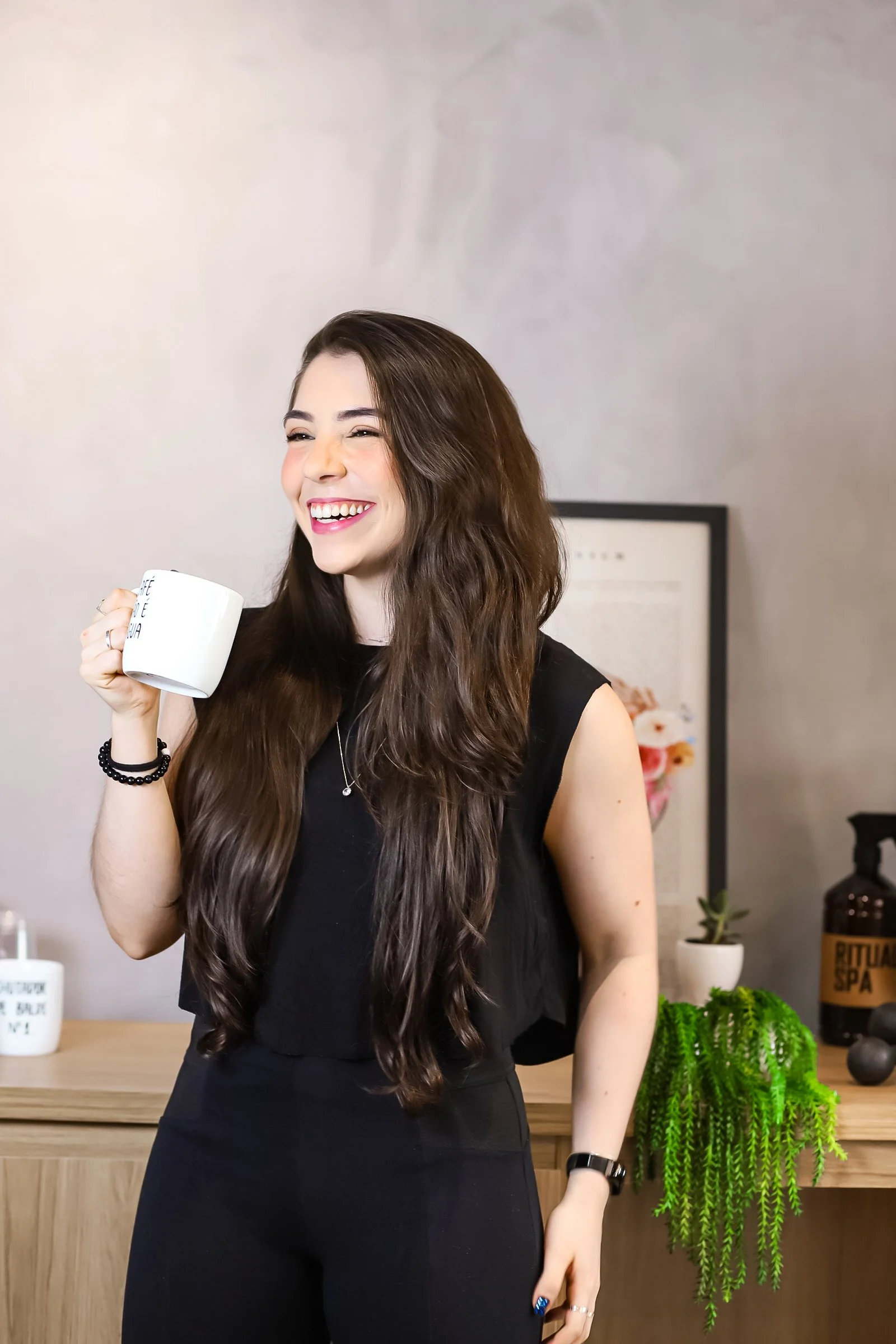 Mulher sorridente com cabelo cacheado escuro, vestindo roupa preta, segurando uma caneca branca em um ambiente interno com decoração simples, incluindo plantas e um quadro ao fundo.