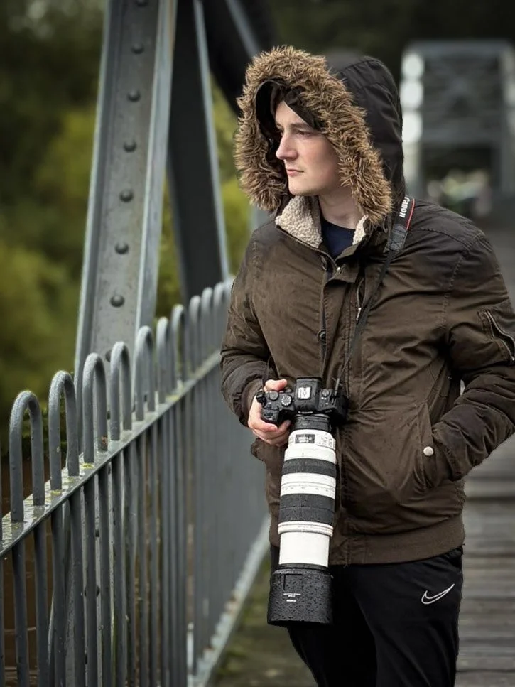 Person with a camera and long zoom lens standing on a bridge, wearing a brown winter coat with a fur-lined hood, looking into the distance.
