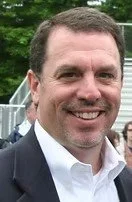 A smiling man with short dark hair, wearing a dark blazer and white shirt, outdoors in a park-like setting.