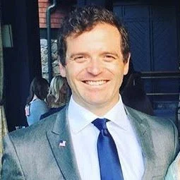 A man in a suit smiling at an outdoor event.