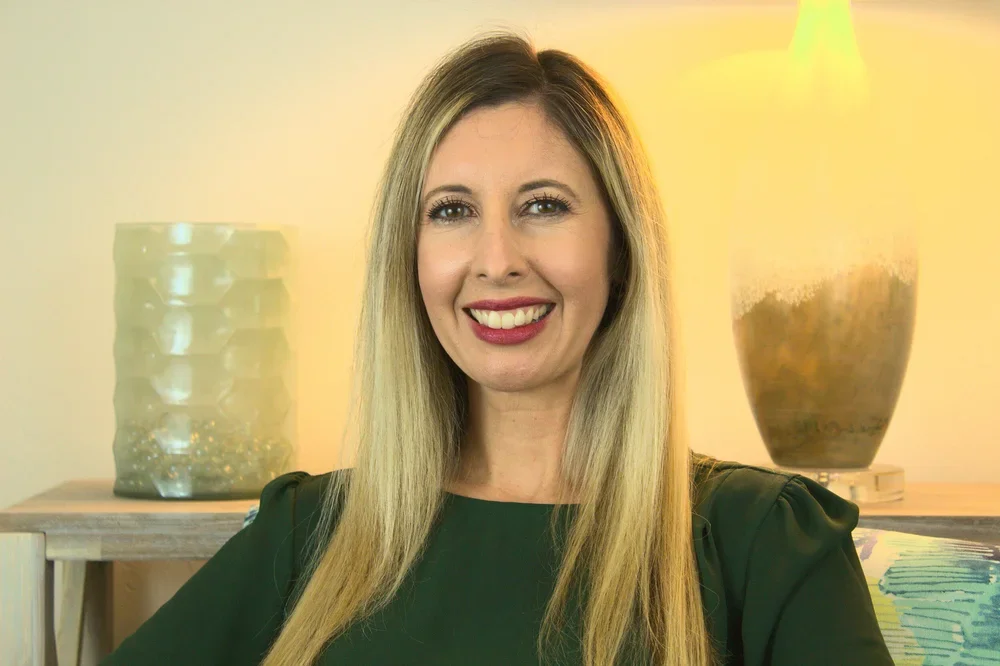 A woman with long blonde hair smiling at the camera in a room with two large vases and warm lighting.