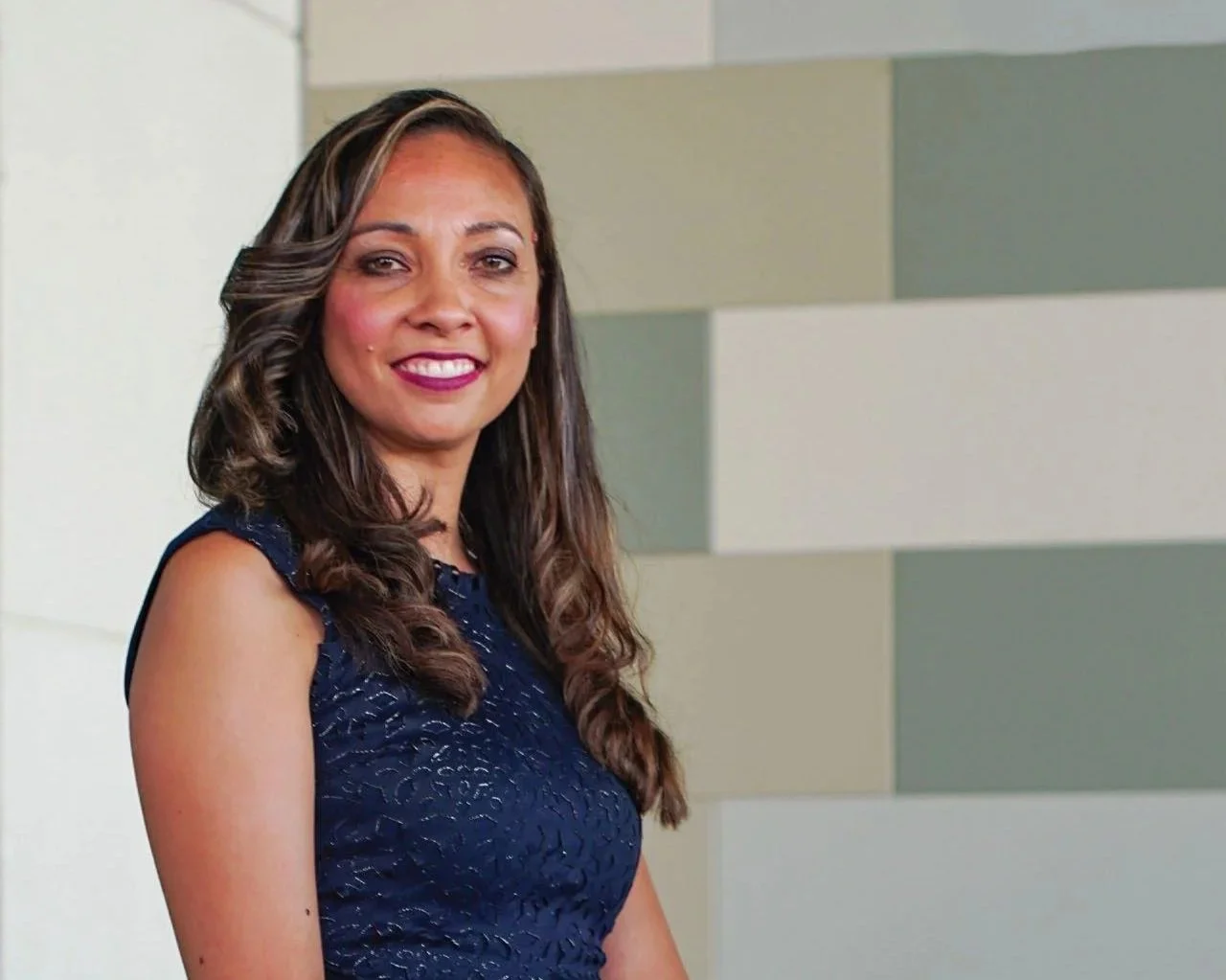 A woman with long, wavy brown hair wearing a navy blue sleeveless dress, smiling in front of a multicolored tiled wall.