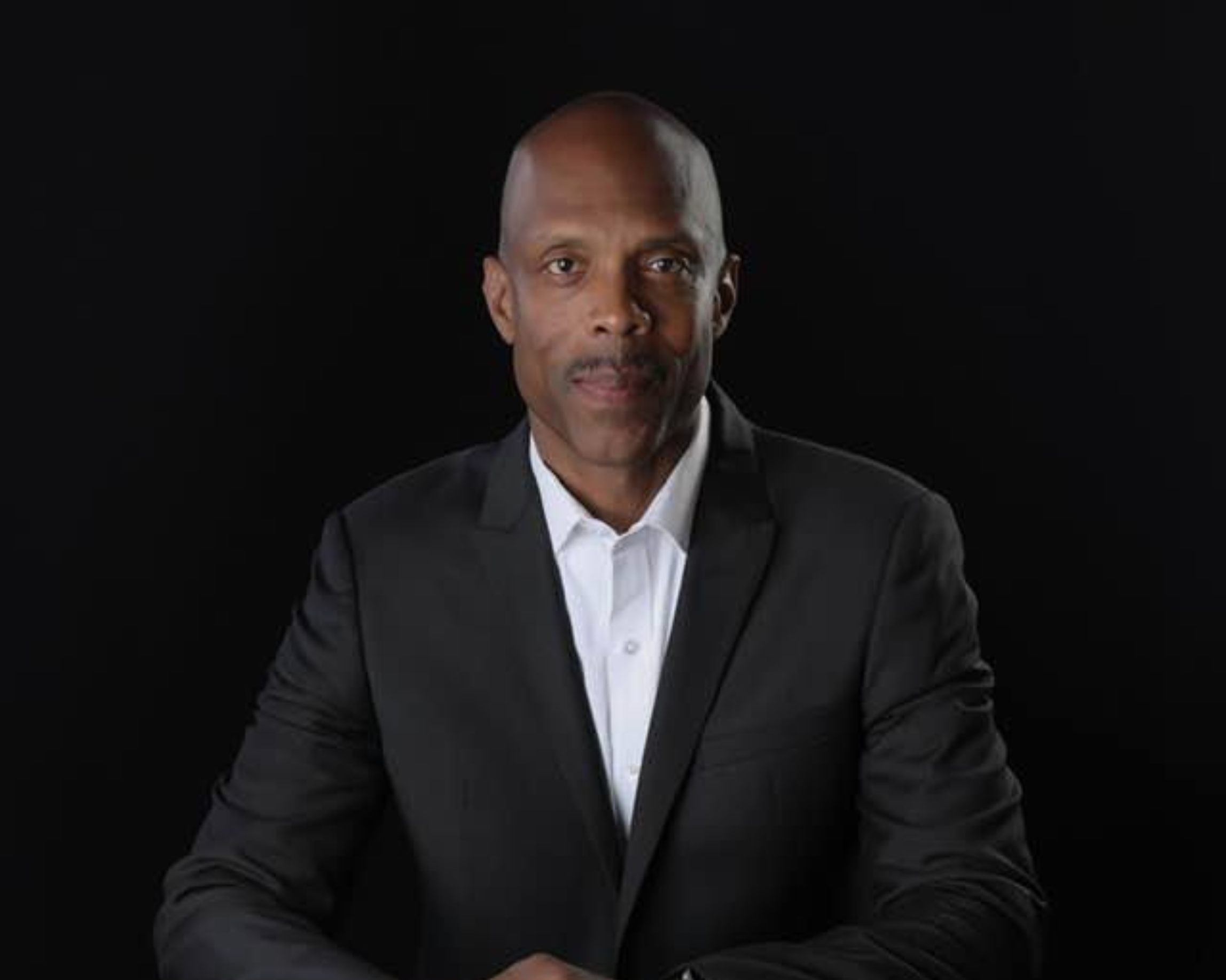 Headshot of a man in a black suit and white shirt against a black background.