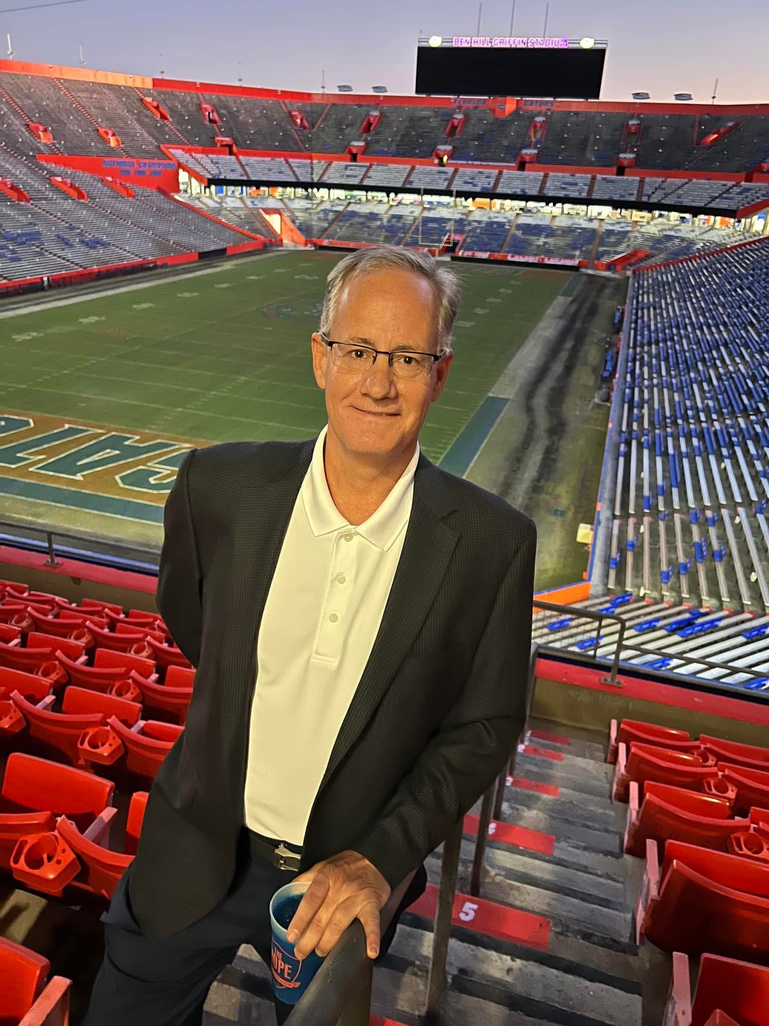 A man in a suit holding a cup stands in a football stadium, with empty red and blue seats and a football field in the background.