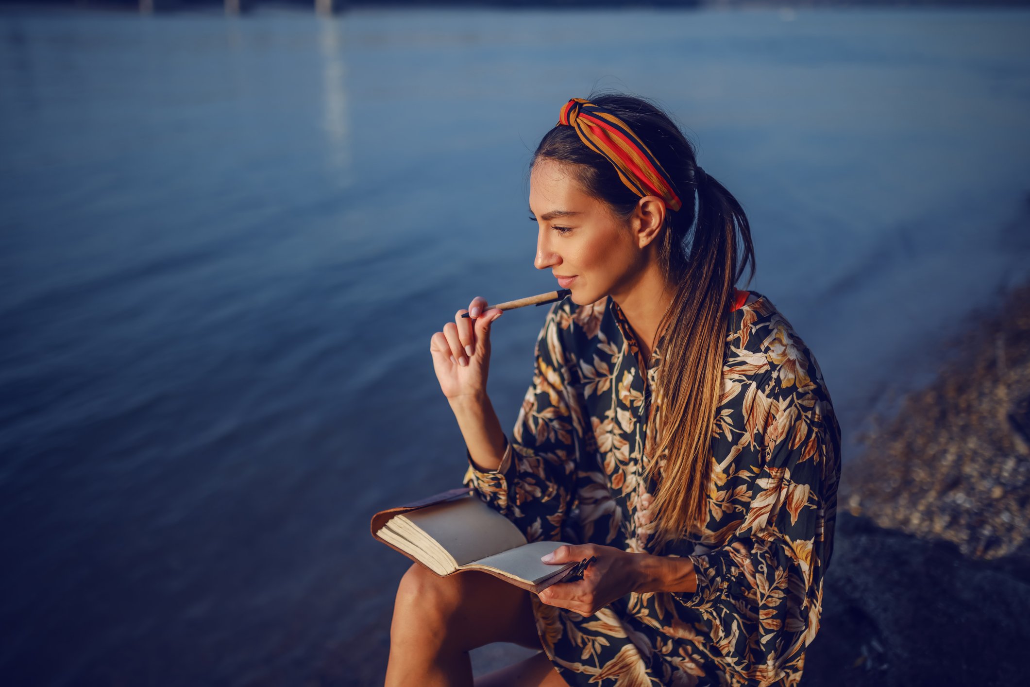 A woman sitting by the water's edge, wearing a floral shirt and a colorful headscarf, holding a book in one hand and a pencil in her mouth while gazing thoughtfully at the water during sunset.