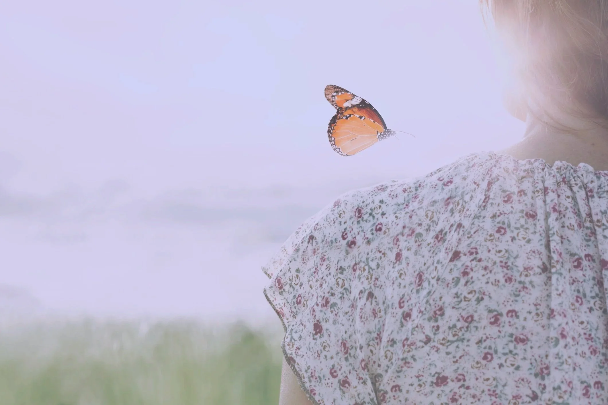 Close-up of a person with blonde hair wearing a floral patterned dress, with an orange and black butterfly resting on their shoulder, outdoors with a blurred background.