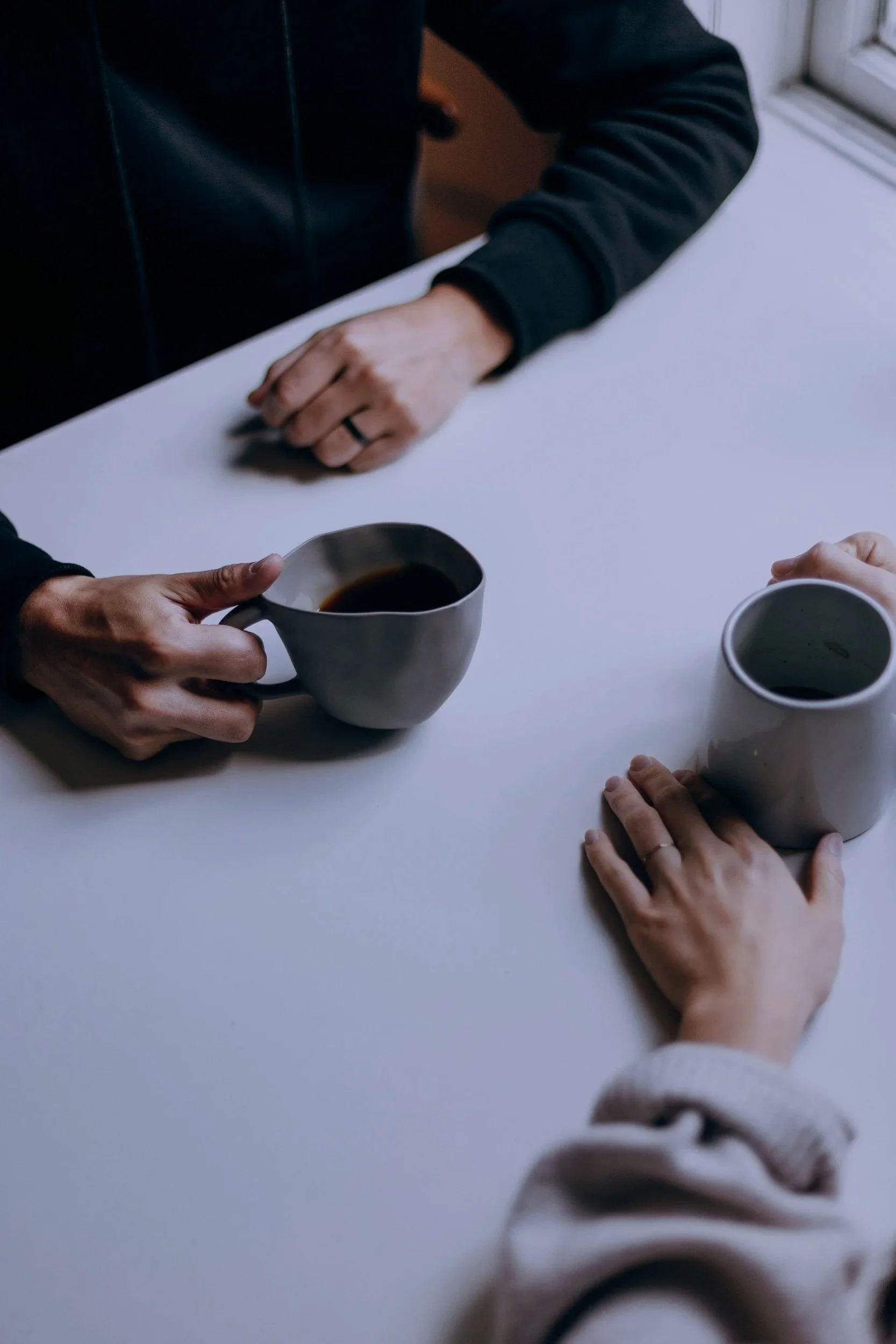 Two people sitting at a white table, each holding a ceramic mug with dark coffee, with one person's hand visible wearing a ring, and the other person's hand resting on the table near a beige sweater.