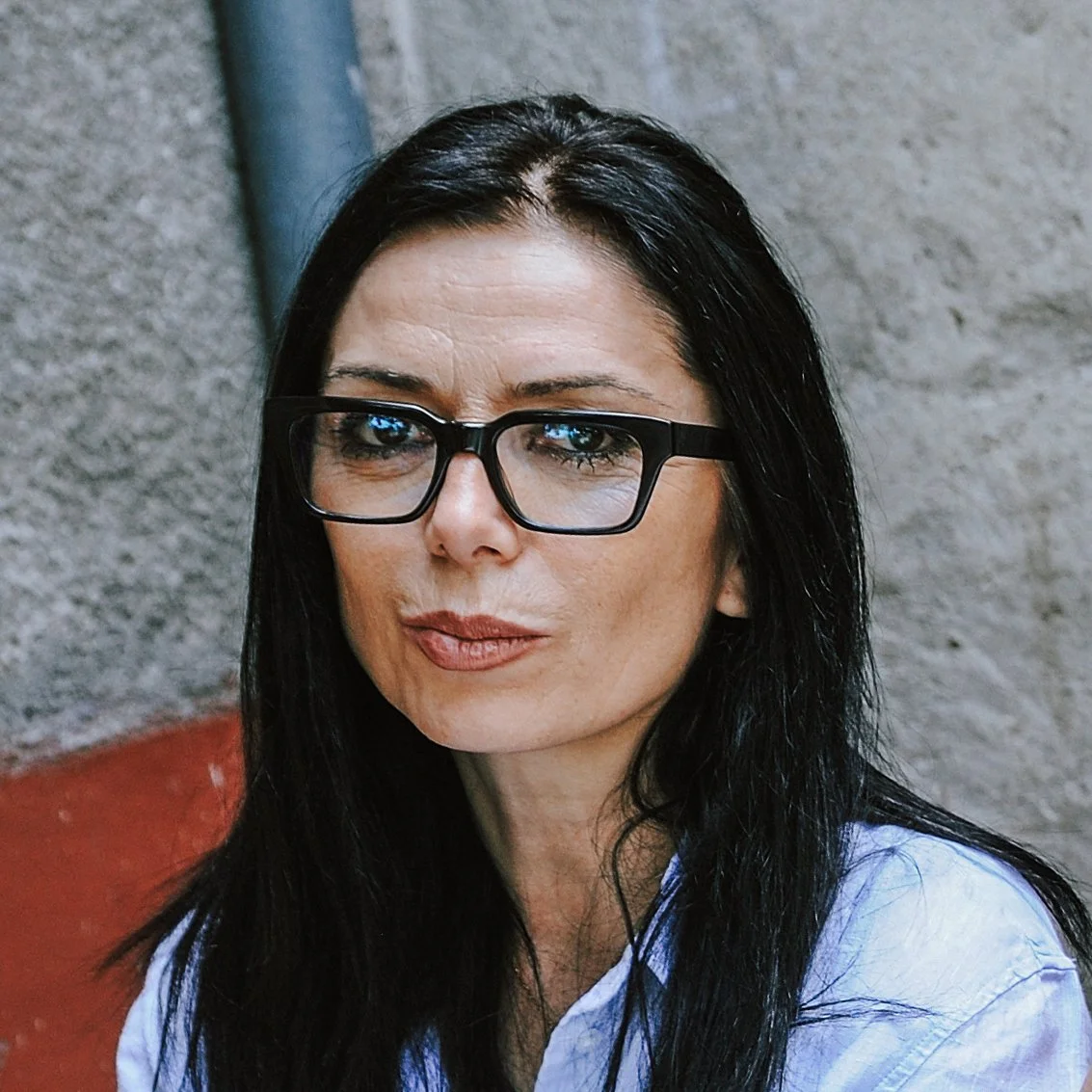 Porträt einer Frau mit schwarzen Haaren und Brille vor einer grauen Wand.