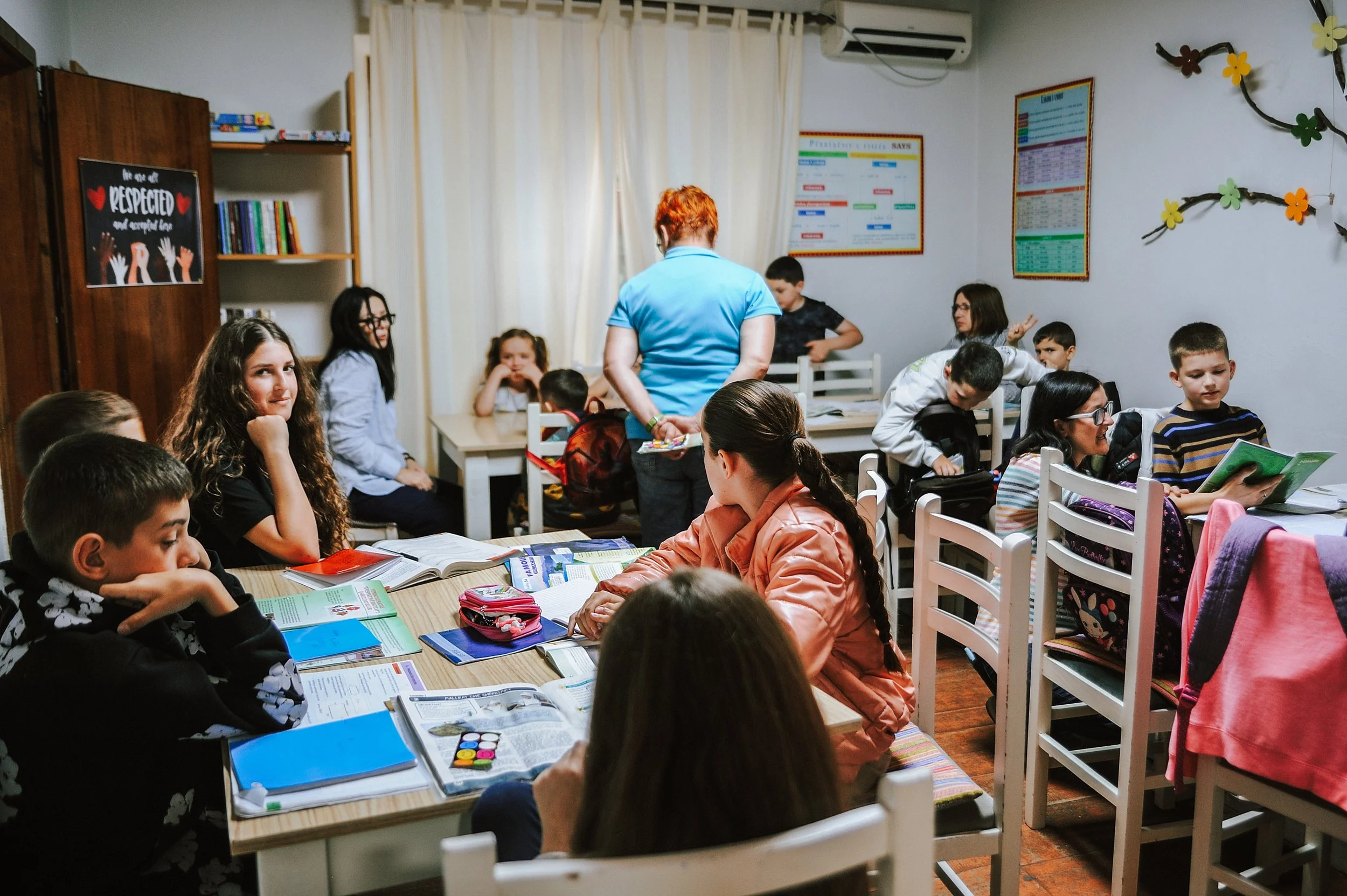 Kindern, die an Tischen im Jungezentrum ASTA sitzen, mit einer Lehrerin, die sich in der Mitte befindet.
