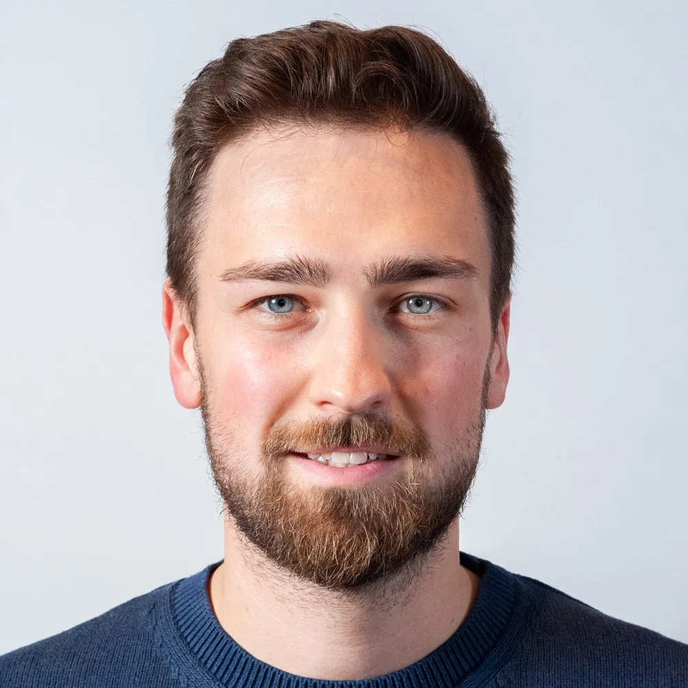 Porträt eines jungen Mannes mit braunem Haar, blauem Blick und Bart, trägt einen blauen Pullover vor einem neutralen Hintergrund.