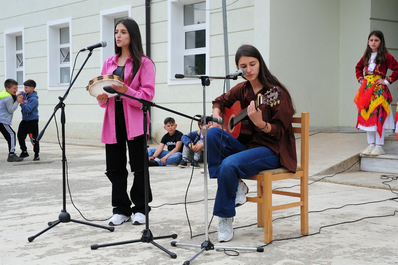 Zwei junge Frauen treten als Musikgruppe auf, eine singt mit Tamburin, die andere spielt Gitarre. Im Hintergrund spielen Kinder, eine jüngere Frau im traditionellen Kostüm steht am rechten Rand auf der Bühne.