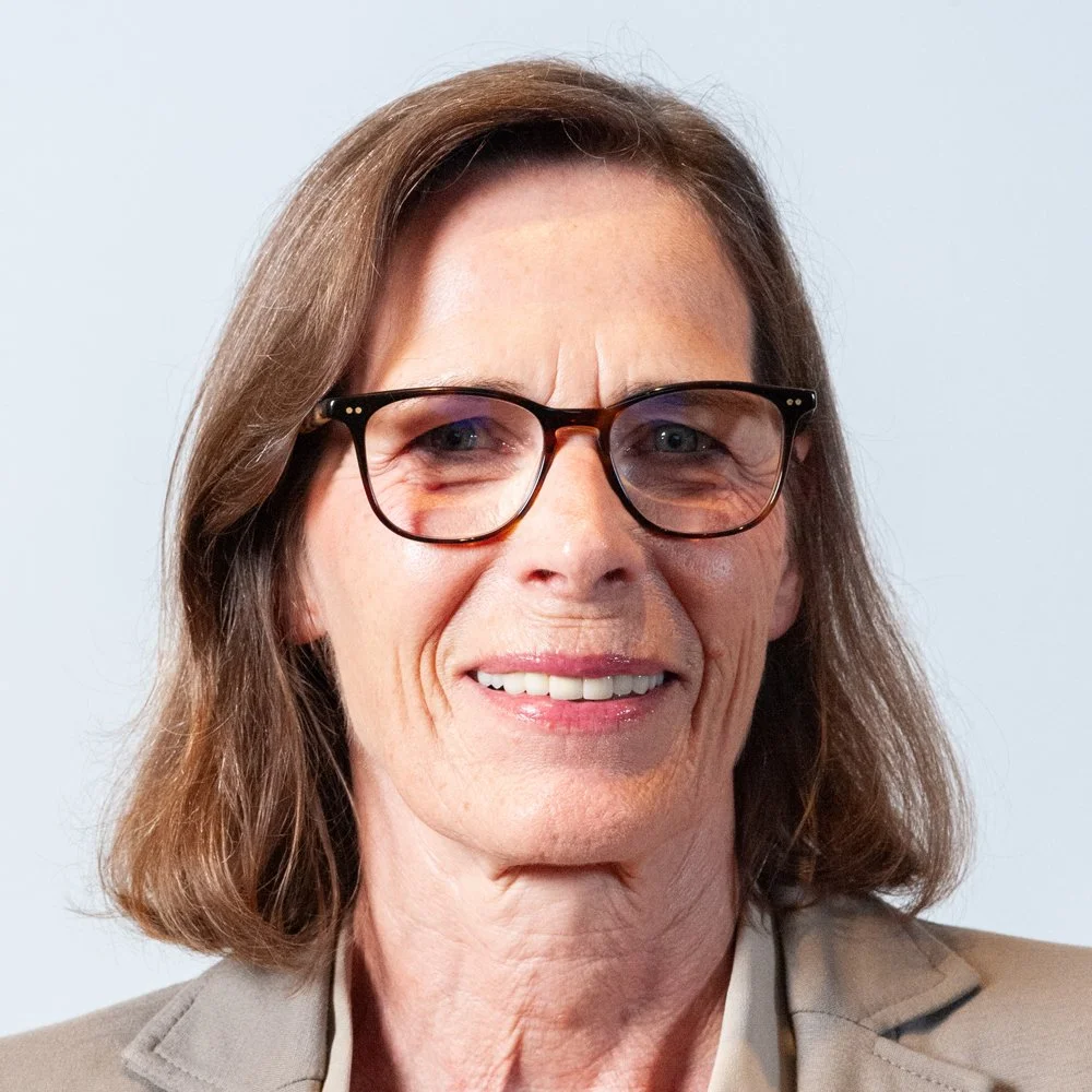 Porträt einer älteren Frau mit Brille und braunen Haaren, lächelnd vor weißem Hintergrund.