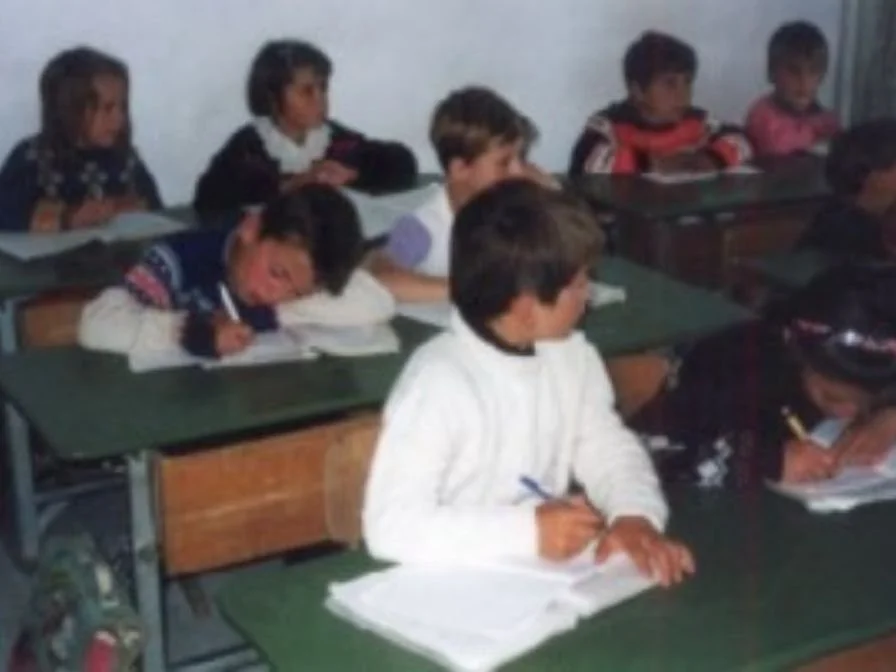 Junge Kinder sitzen in einer Schulklasse, schreiben in Hefte, einige schauen nach vorne, während andere Notizen machen.