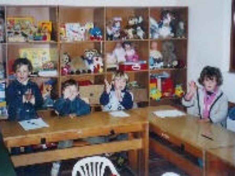Vier Kinder sitzen an Tischen in einem Raum mit Holzregalen im Hintergrund, die Spielzeuge und Stofftiere enthalten.