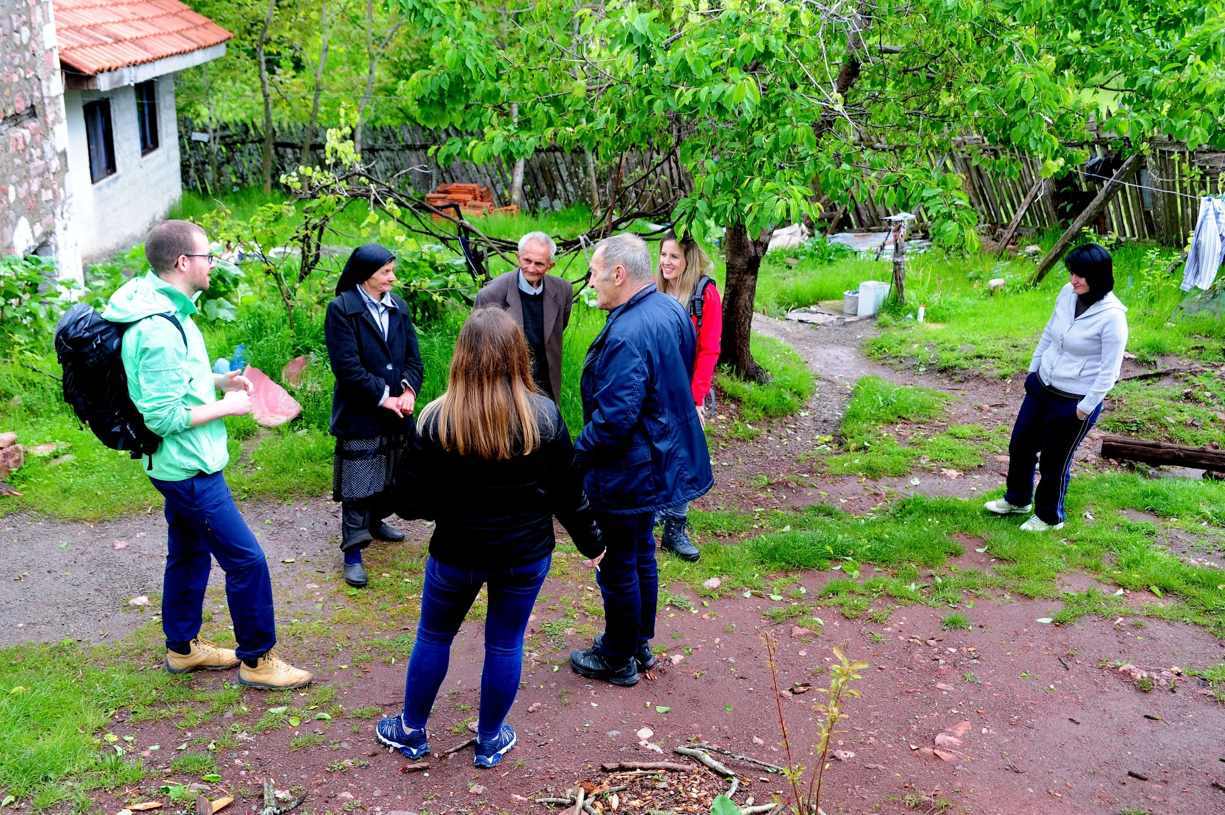 Eine Gruppe von Menschen, u.a. Männer und Frauen, stehen im Garten unter einem Baum und sprechen miteinander. Einige tragen Jacken und Rucksäcke, eine Frau hat dunkle Haare und trägt eine hellgraue Jacke, ein Mann hat kürzere Haare und trägt eine dunkle Jacke. Es ist eine sonnige Tageszeit, umgeben von grünem Gras, Bäumen und einem alten Holzzaun.