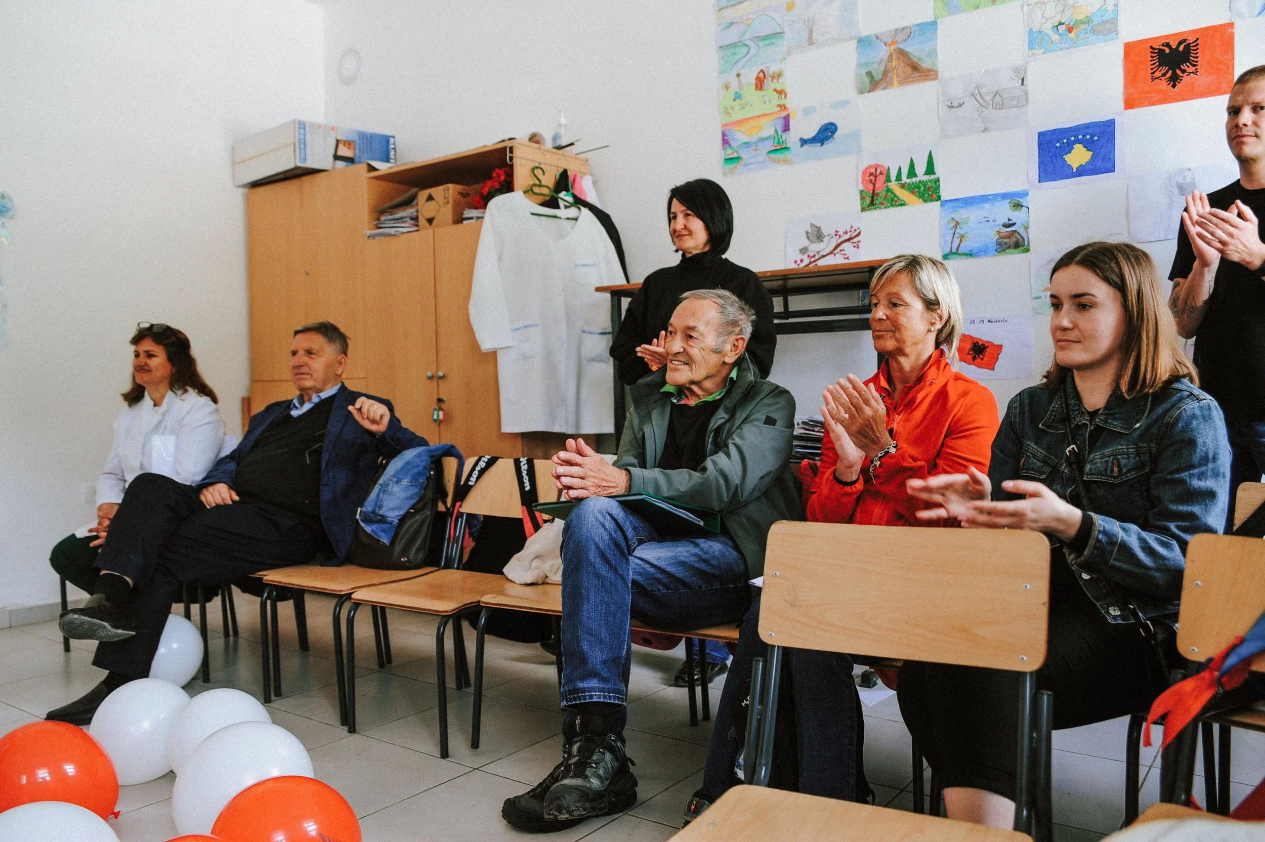 Menschen sitzen in einem Raum, applaudieren und schauen nach vorne. Das Klassenzimmer ist mit Kinderzeichnungen an der Wand dekoriert, und es gibt Ballons auf dem Boden.