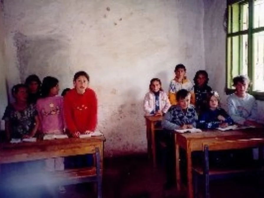 Kinder in einer Schulklasse sitzen an Holztischen, eine Gruppe steht an einem Tisch, eine Gruppe sitzt durch einen Fensterrahmen auf der rechten Seite, die Wand im Hintergrund ist ungleichmäßig und schlicht.